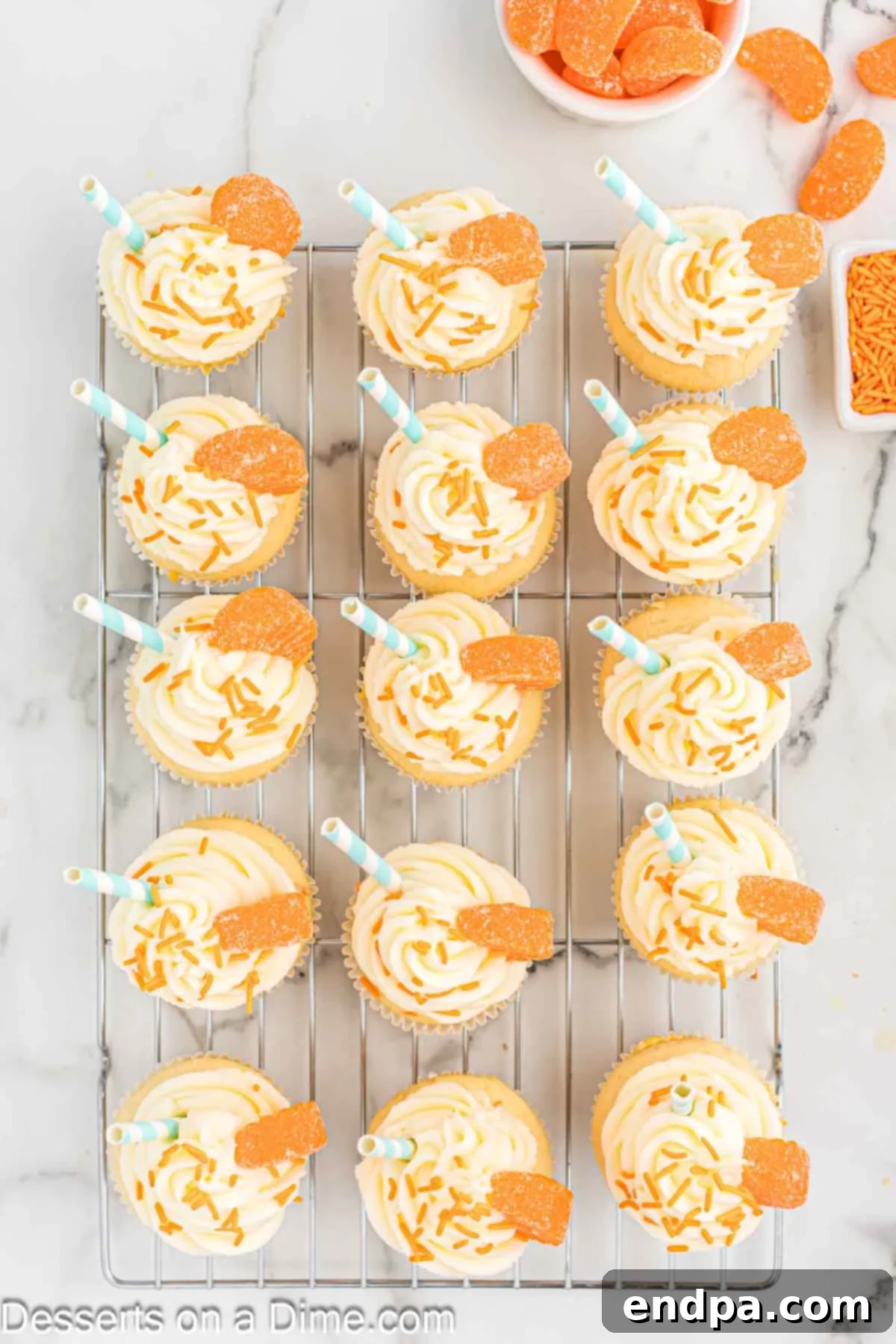 Fully frosted orange creamsicle cupcakes on a wire rack, decorated with orange sprinkles, candied orange slices, and small paper straws, ready to be served.