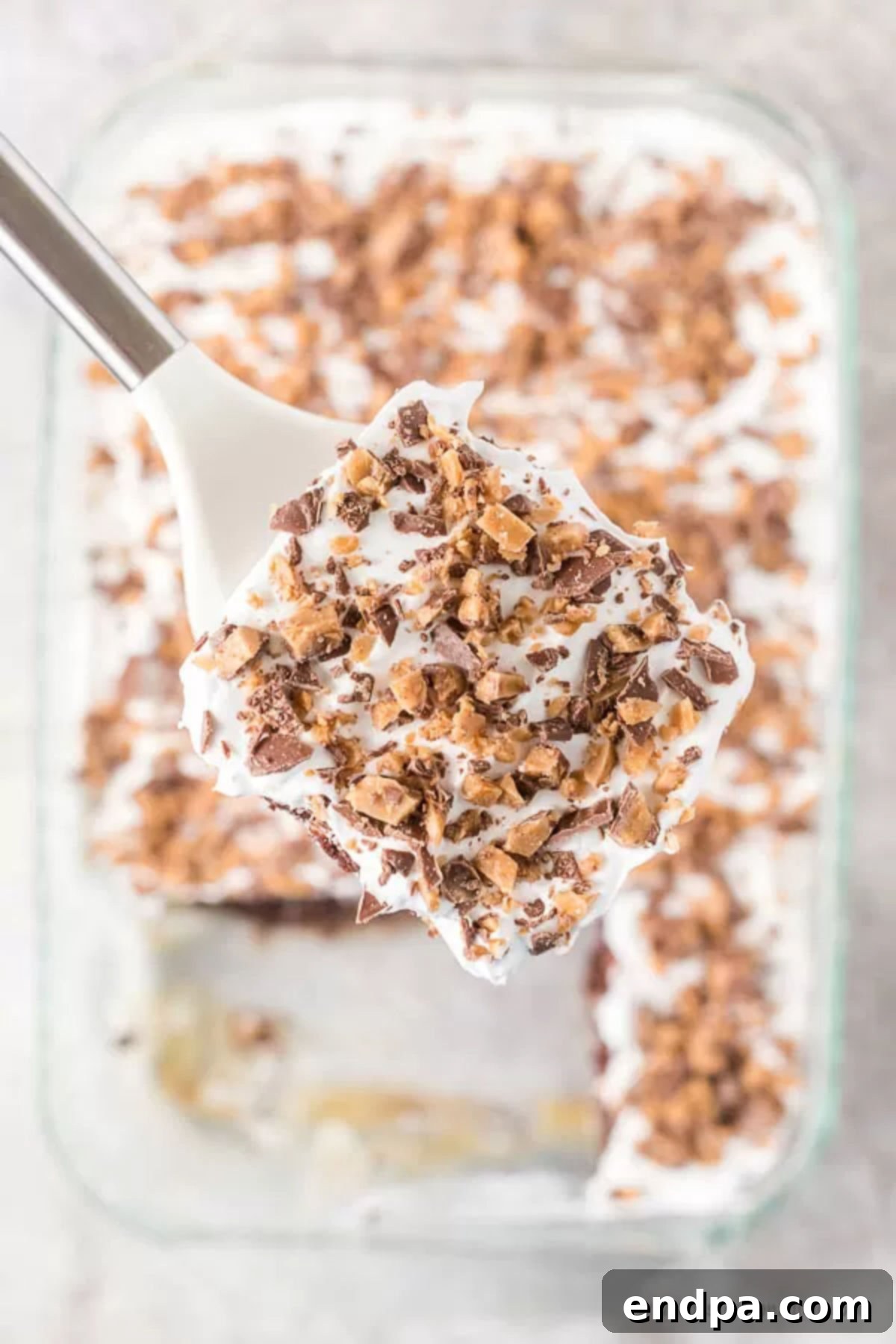 Better than sex cake recipe is so decadent! It's also known as heath bar cake recipe and each bite is loaded with caramel, chocolate, toffee and more. Heath bar cake recipe desserts are so easy and chocolate heath bar cake is a must try. Heath bar cake condensed milk and other simple ingredients make such an easy heath bar cake. #dessertonadime #betterthansexcakerecipe A close up image of a slice of better than sex cake on a spatula with a whole cake in the background