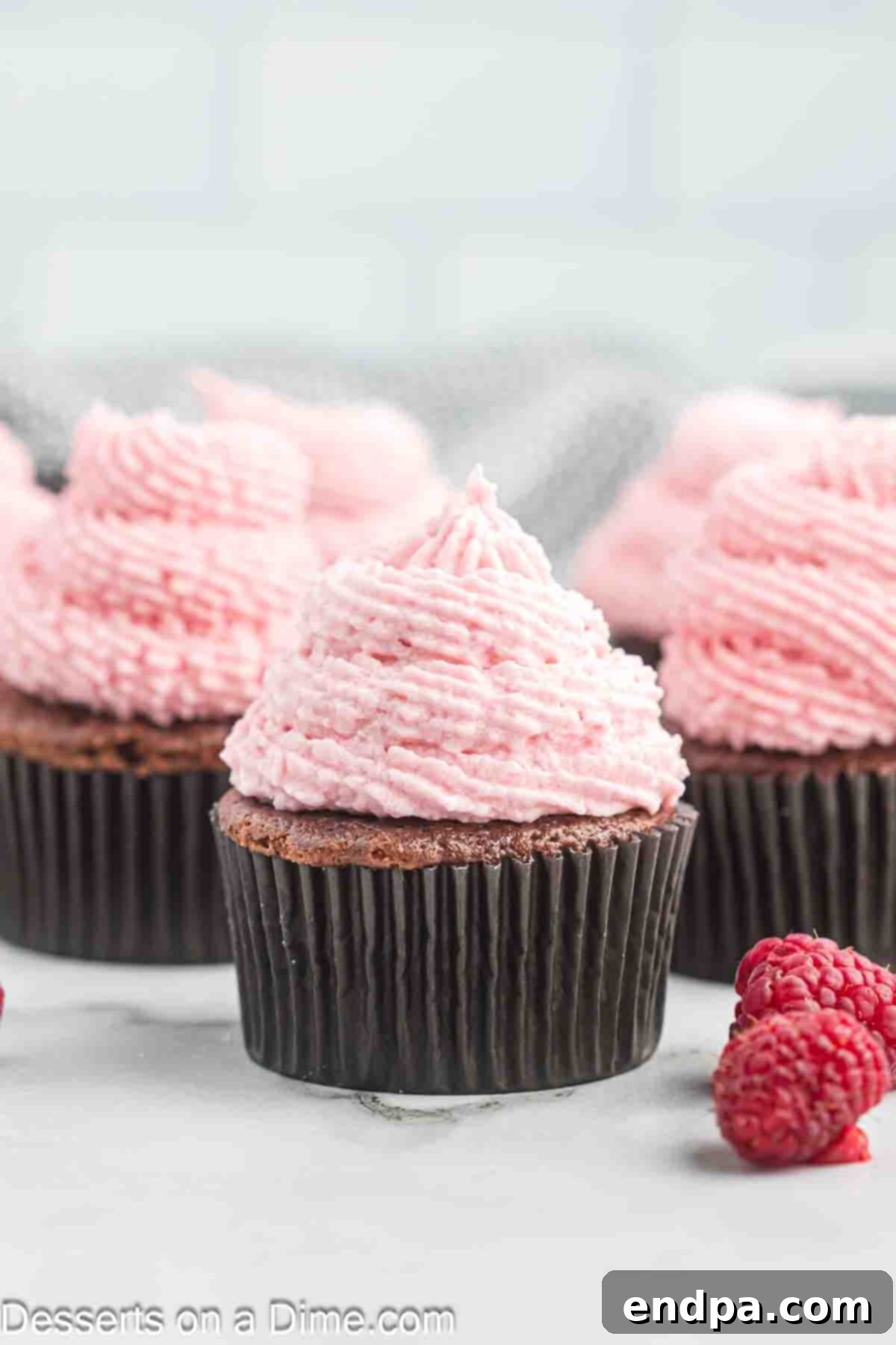 Two cupcakes topped with beautiful raspberry frosting and fresh raspberries, against a blurred background.