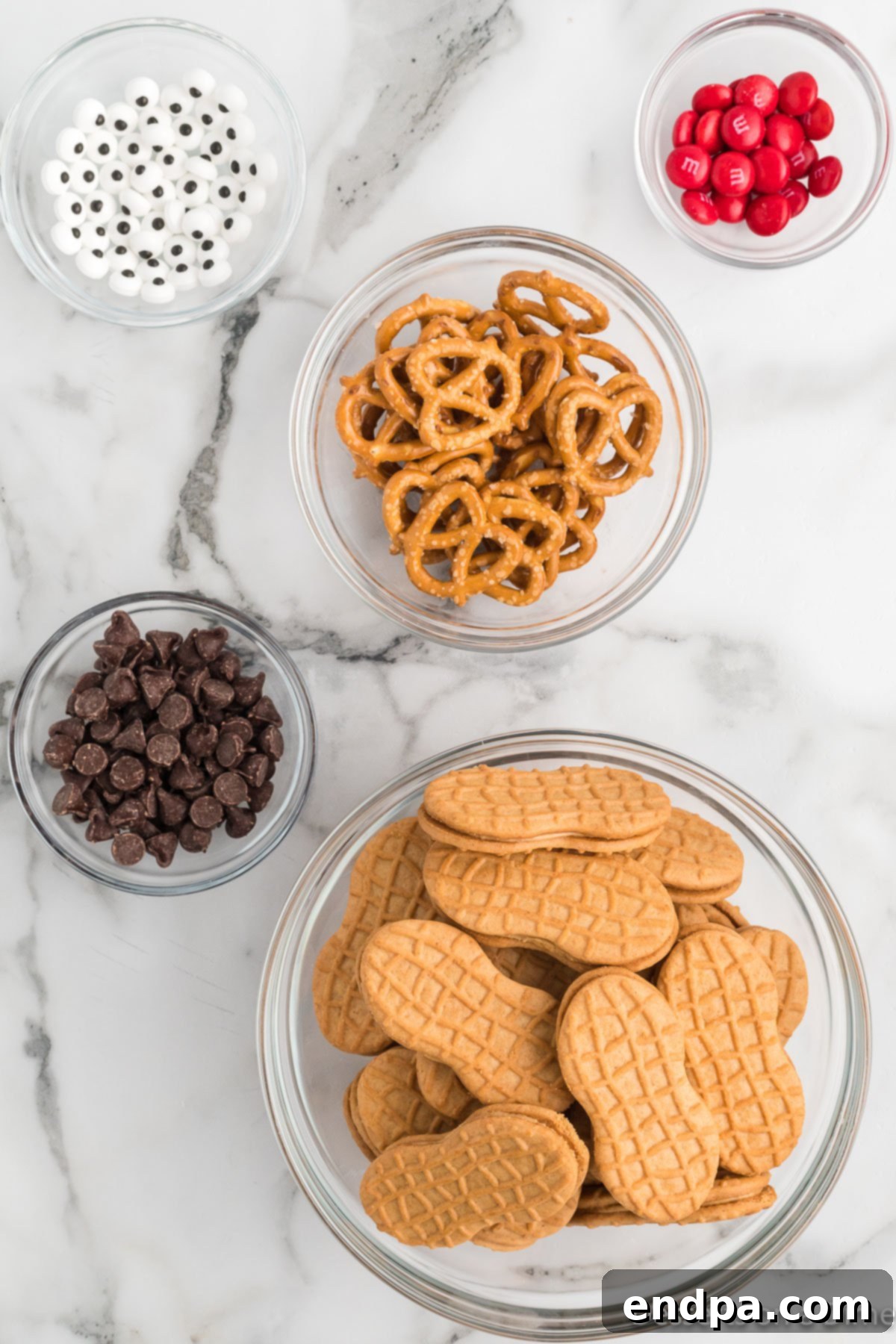 A selection of ingredients laid out, including Nutter Butter cookies, mini pretzels, candy eyes, red M&Ms, and chocolate chips.