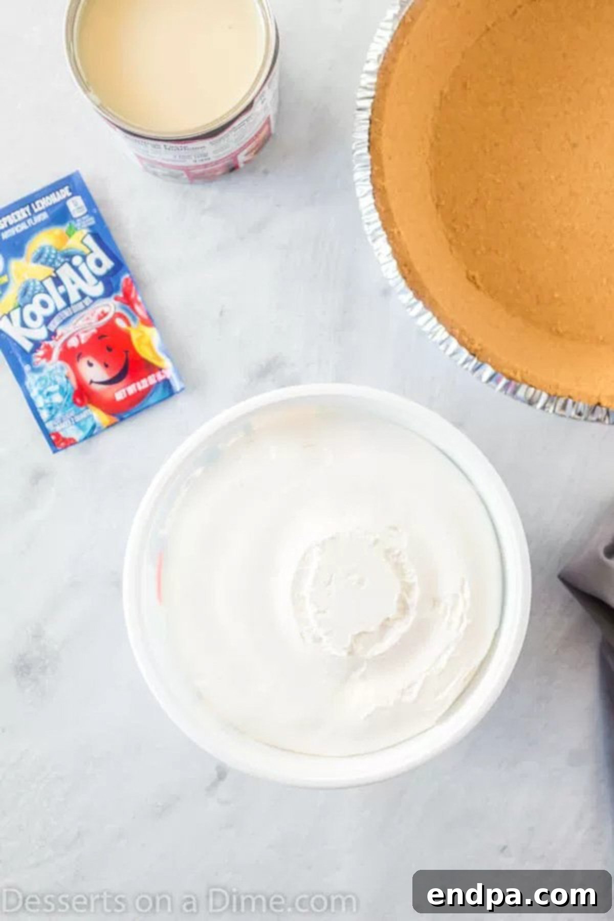 The simple ingredients for a Kool-Aid pie laid out: a package of blue Kool-Aid mix, a can of sweetened condensed milk, a container of whipped topping, and a pre-made graham cracker pie crust.