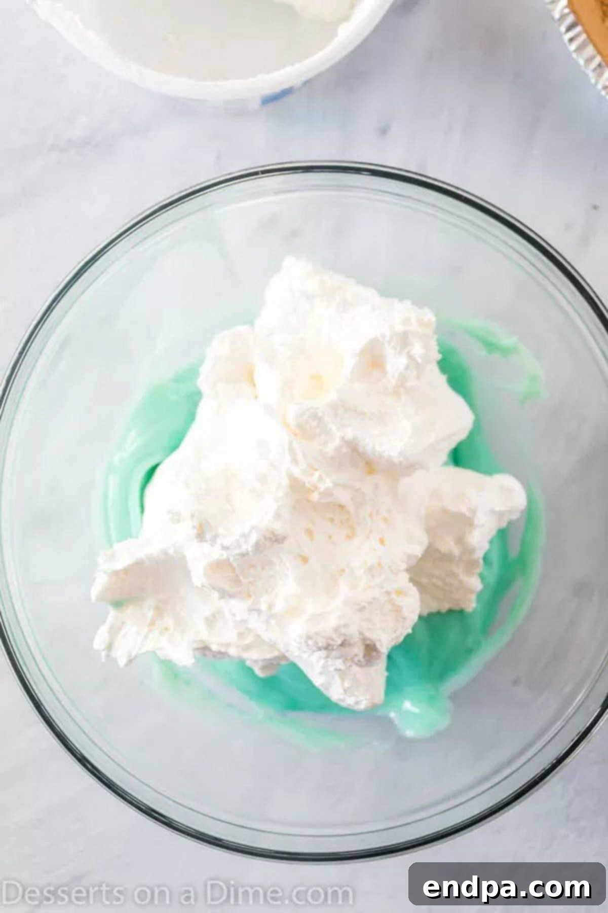 A mixing bowl filled with a creamy, colorful Kool-Aid pie filling, topped with a dollop of whipped cream before being folded in.