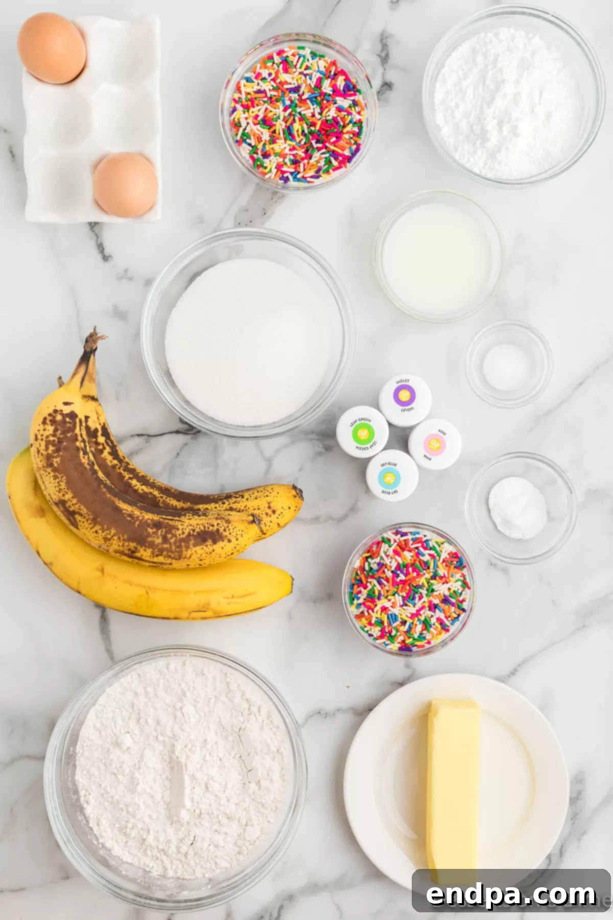 Ingredients for Unicorn Bread: flour, baking soda, sugar, butter, eggs, bananas, food coloring, and sprinkles, laid out on a surface.