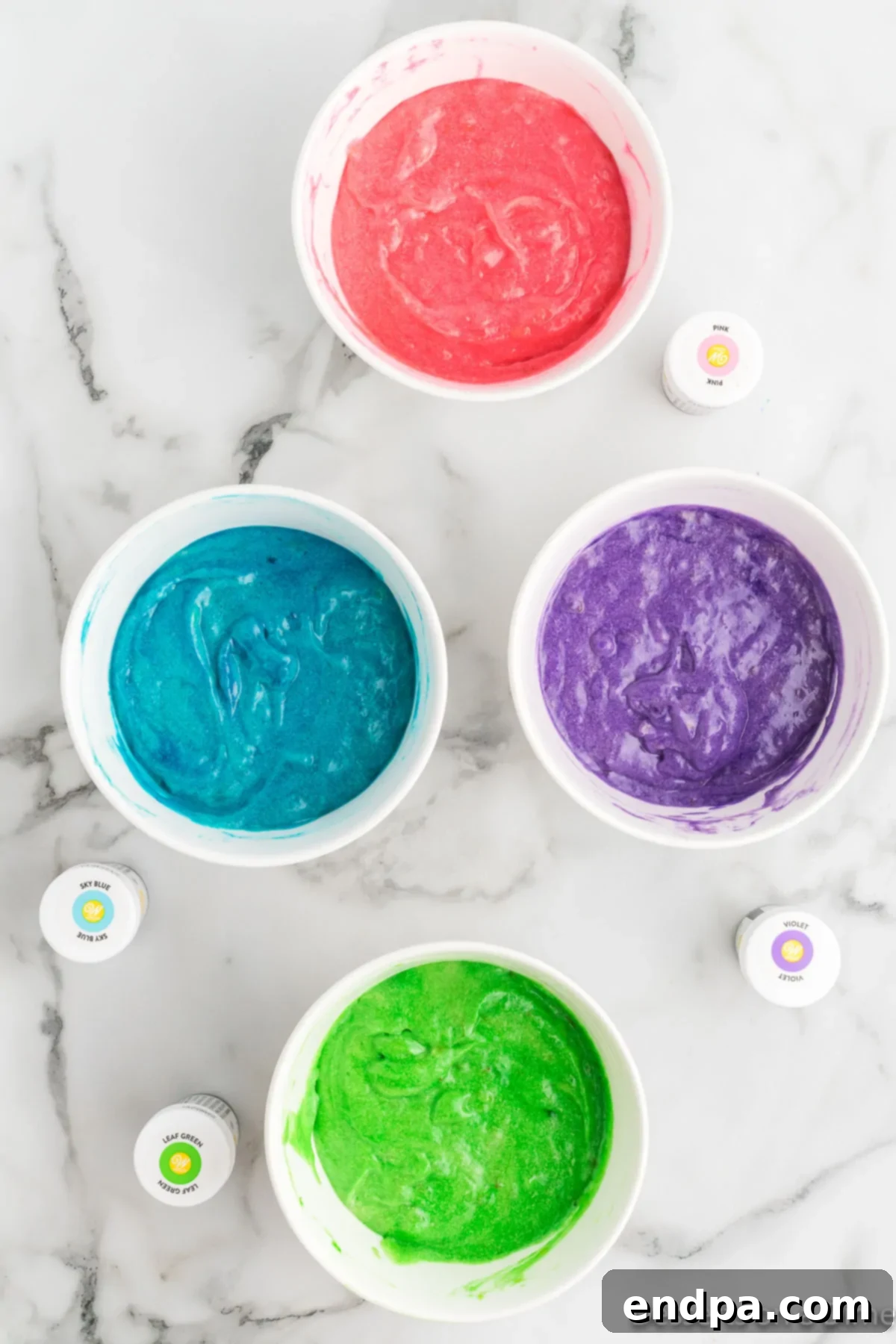 Four small bowls containing batter dyed with different food colors: pink, green, purple, and blue.