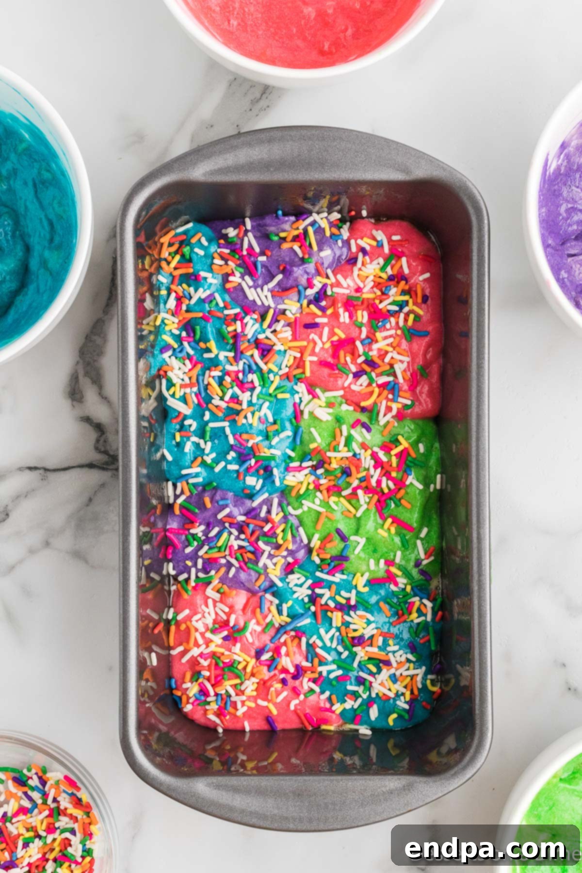 Layered colorful batter in a loaf pan, topped with sprinkles, ready for baking.