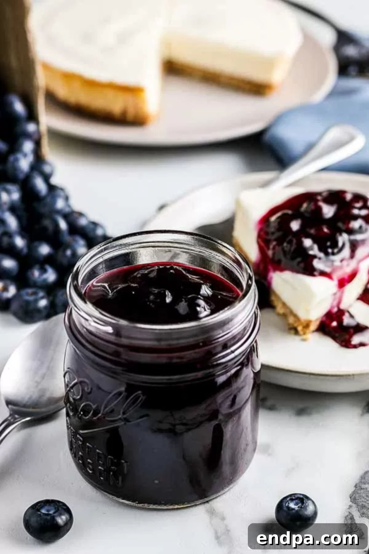 Close up image of blueberry sauce in a mason jar with a slice of cheesecake with blueberry sauce on top