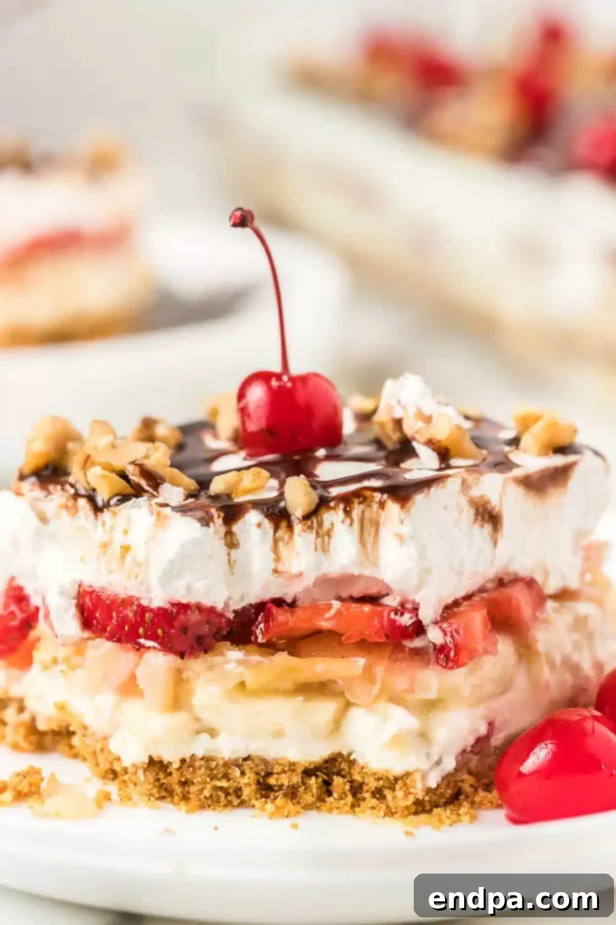 Easy No-Bake Banana Split Cake is a hit. Layers of strawberries, crushed pineapple, bananas, and a creamy filling topped with chocolate and cherries make it a decadent no-bake dessert. A beautiful slice of No Bake Banana Split Cake on a white plate, showcasing its vibrant layers of fruit, cream, and chocolate drizzle.