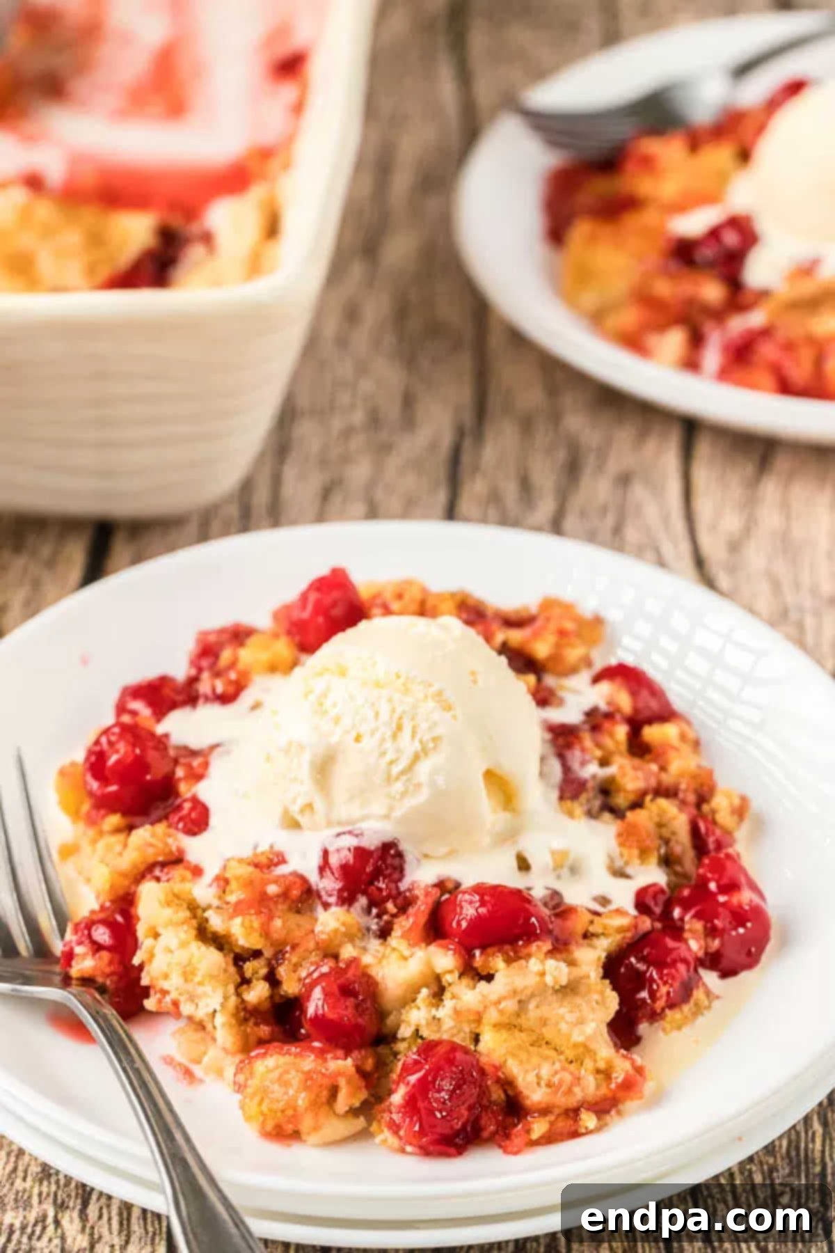 A slice of warm cherry dump cake on a white plate, topped with a melting scoop of vanilla ice cream.