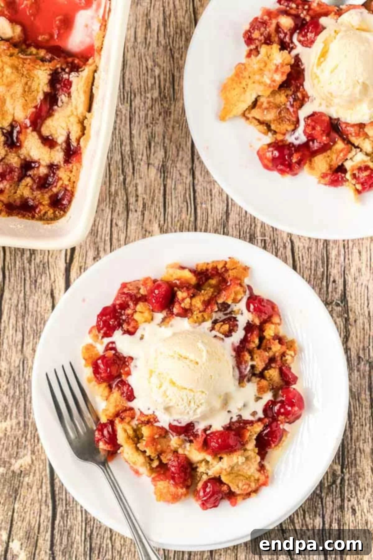 A close-up of a slice of cherry dump cake on a plate, with a fork digging into the warm cake and a scoop of vanilla ice cream.