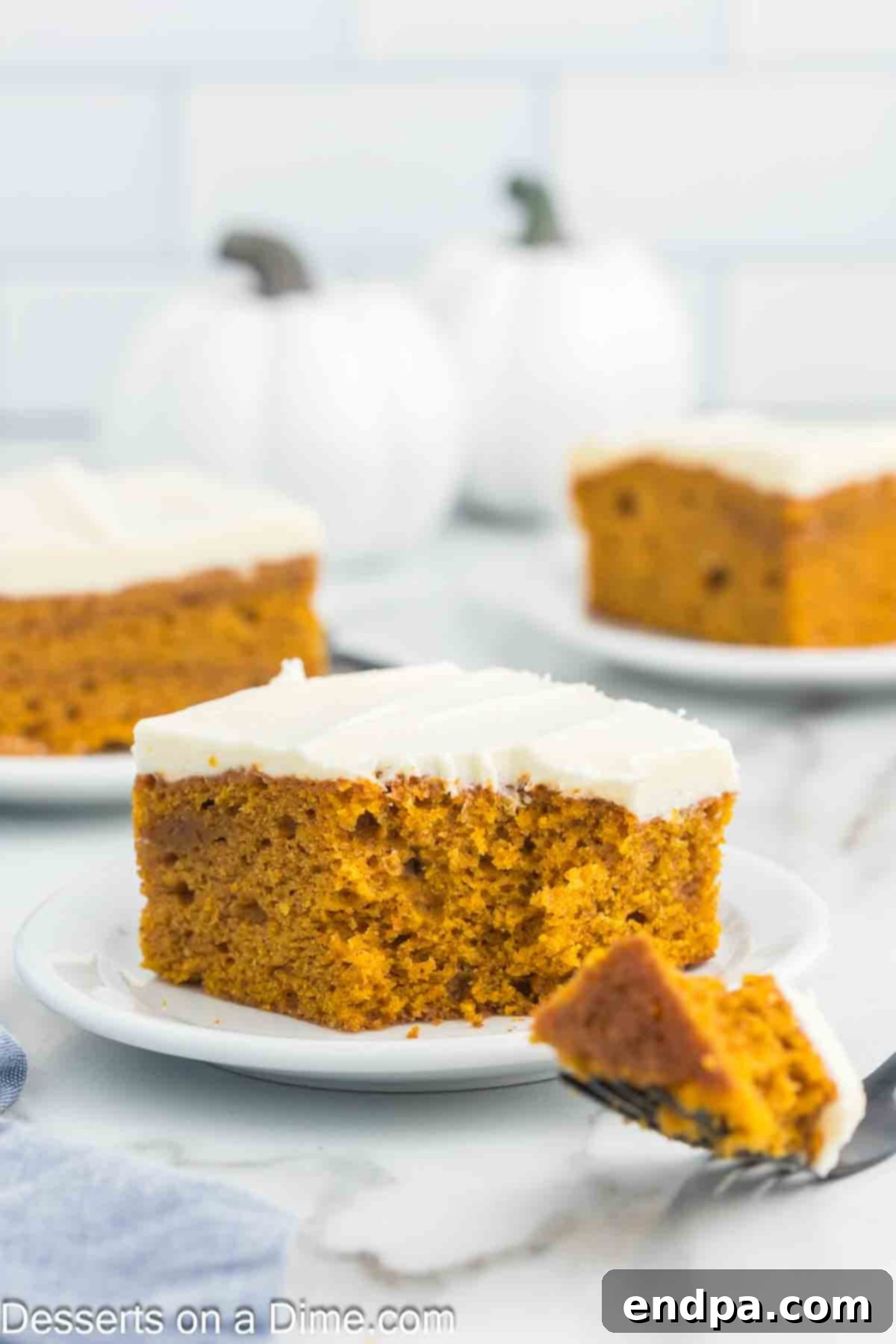 A beautifully presented slice of incredibly moist Pumpkin Cake, generously topped with a thick layer of creamy, white cream cheese frosting, resting on a pristine white plate, garnished with a sprinkle of cinnamon. The background suggests a warm, inviting autumn setting.