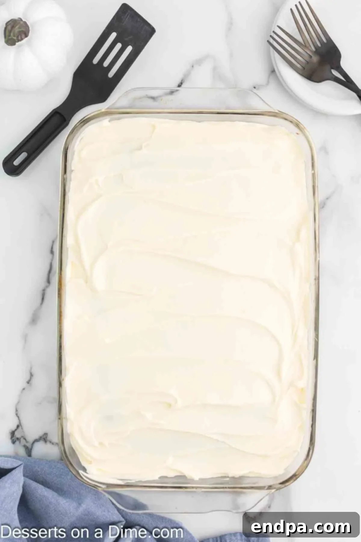 The completely cooled pumpkin cake is being lavishly frosted with the freshly prepared homemade cream cheese frosting. A spatula is used to spread the rich, white frosting evenly and artfully across the entire surface of the cake, creating inviting swirls.