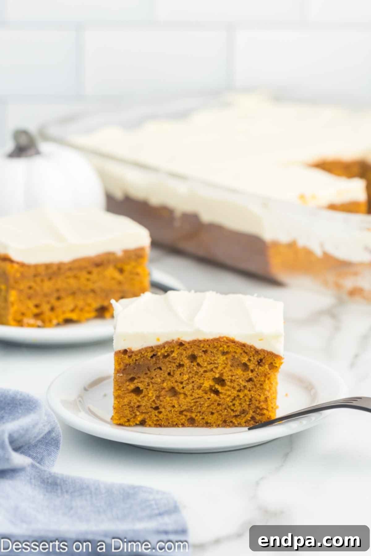 A perfectly portioned slice of golden pumpkin cake, layered with a thick, smooth application of white cream cheese frosting, is beautifully presented on a clean white plate. The cake shows its moist, tender crumb texture, ready to be enjoyed.