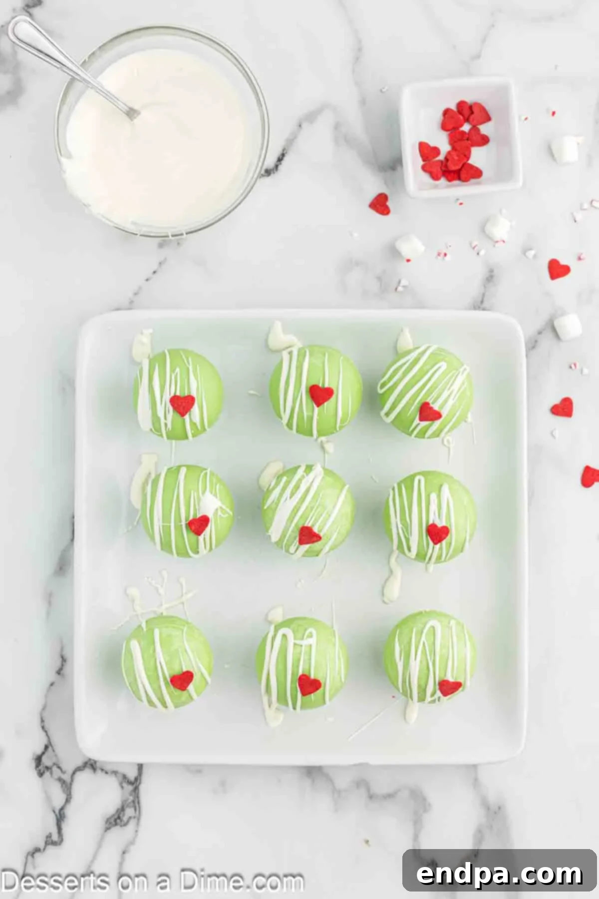 Grinchmas Hot Cocoa Bombs 8 Grinch hot chocolate bombs being drizzled with melted white candy melts and topped with a red heart sprinkle.
