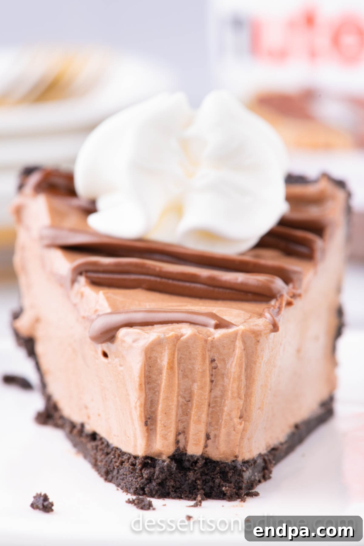 Decadent No Bake Nutella Cheesecake 12 A close-up of a slice of No Bake Nutella Cheesecake on a white plate, showcasing the rich, dark filling, distinct Oreo crust, and a decorative drizzle of Nutella on top.