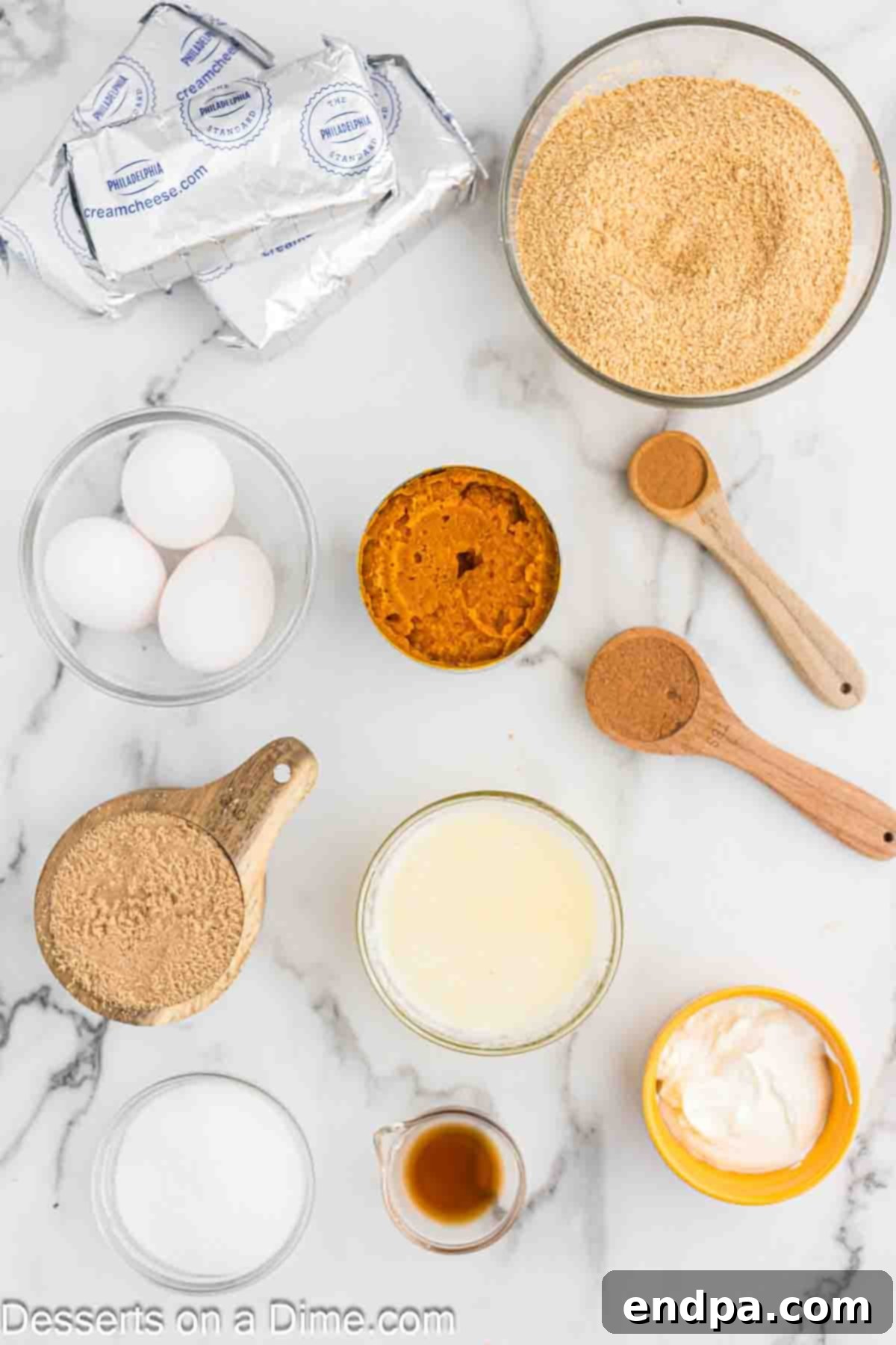 Velvet Pumpkin Spice Cheesecake 3 A collection of fresh ingredients laid out on a wooden surface for pumpkin cheesecake, including graham cracker crumbs, cubes of butter, granulated sugar, a jar of cinnamon, blocks of cream cheese, a can of pure pumpkin puree, fresh eggs, a container of sour cream, a bottle of vanilla extract, and a small jar of pumpkin pie spice.