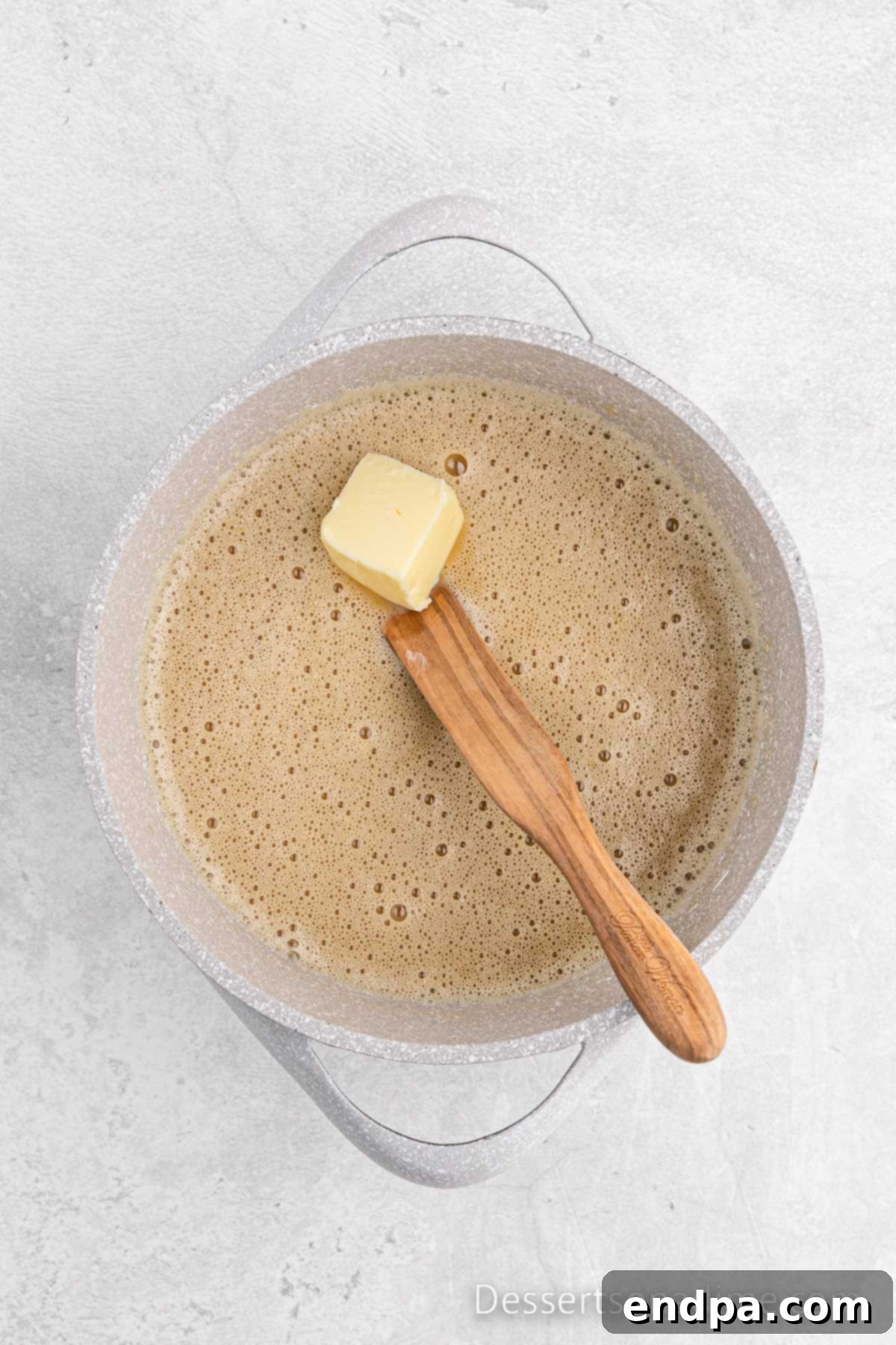 Golden Butterscotch Delight 5 Two tablespoons of unsalted butter, cut into small pieces, are being melted into the thickening butterscotch mixture in a saucepan, being constantly stirred with a whisk to ensure a smooth, glossy, and fully incorporated finish for the pie filling.