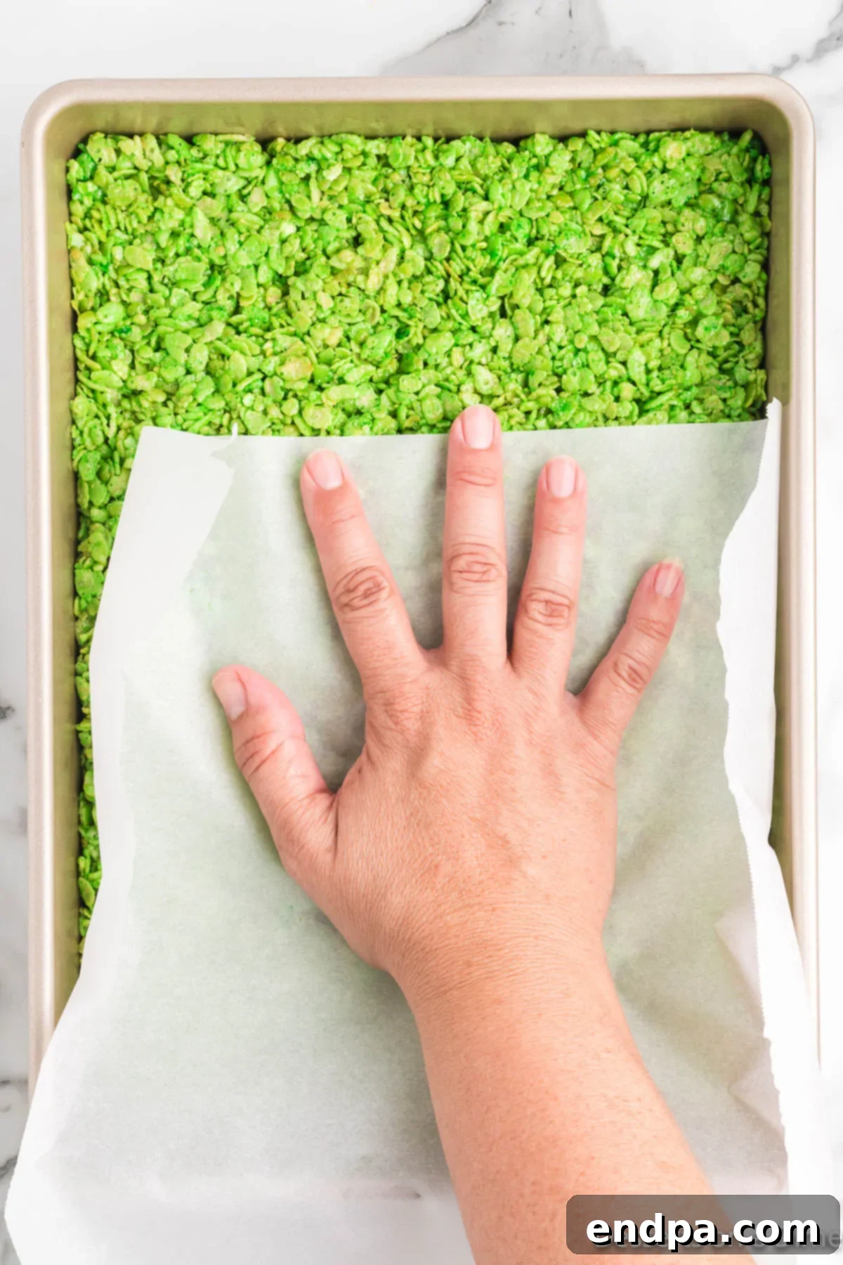Green Rice Krispie treat mixture being pressed evenly into a prepared 9x13 inch baking pan.