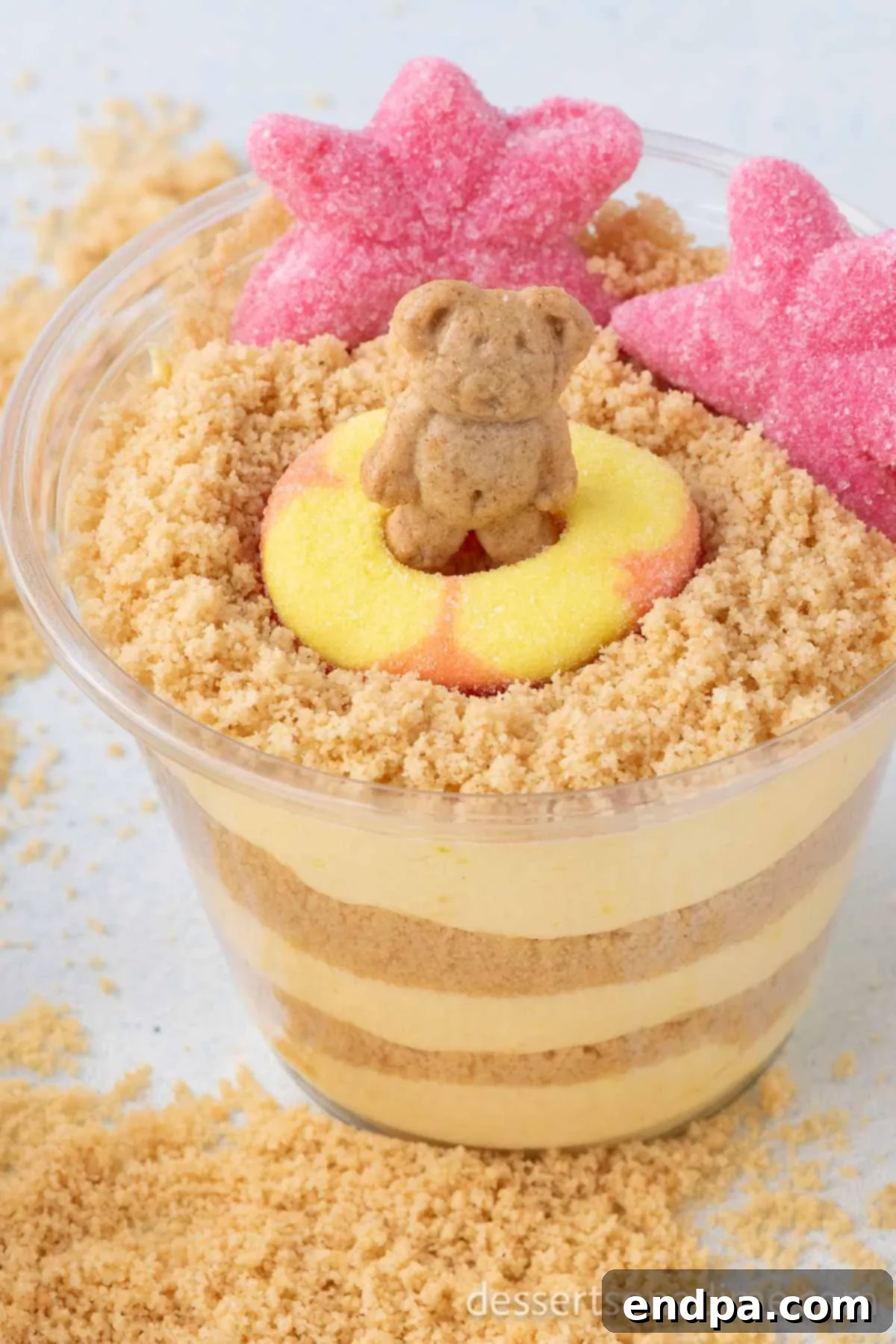 Delicious Sand Pudding Cups presented in clear plastic cups, ready to be enjoyed at a summer party. The top layer of crushed cookies resembles sand, with gummy candies and teddy grahams creating a fun beach scene.