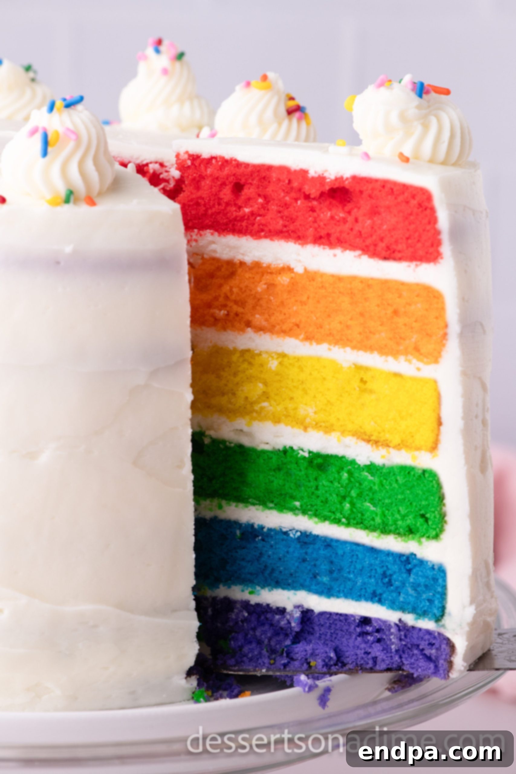 Kaleidoscope Delight 2 A slice of Rainbow Cake revealing its six vibrant, colorful layers and fluffy buttercream frosting, artfully presented on a white plate, highlighting its moist texture.