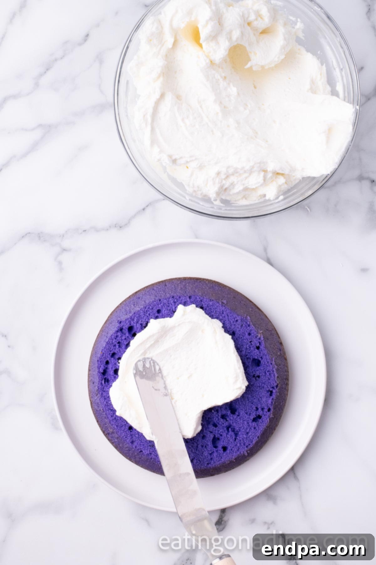 Kaleidoscope Delight 9 The first purple layer of the Rainbow Cake placed on a large plate, with a generous, even layer of white buttercream frosting spread on top, ready for the next colorful layer.