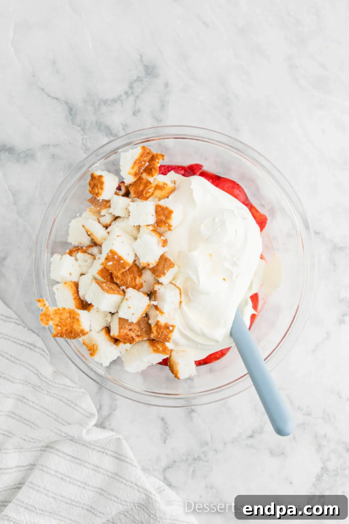 Diced angel food cake cubes being gently folded into the strawberry Jell-O and Cool Whip mixture in a large bowl.