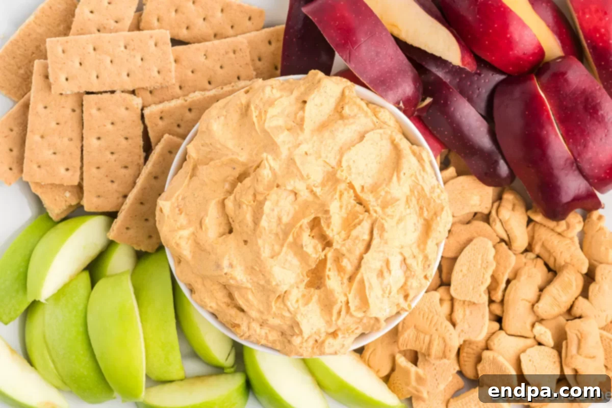 Autumn Pumpkin Fluff Dip 2 A bowl of creamy Pumpkin Dip with Cool Whip, garnished with a sprinkle of cinnamon, surrounded by an assortment of dippers including apple slices, graham crackers, and vanilla wafers, set against a cozy fall backdrop.