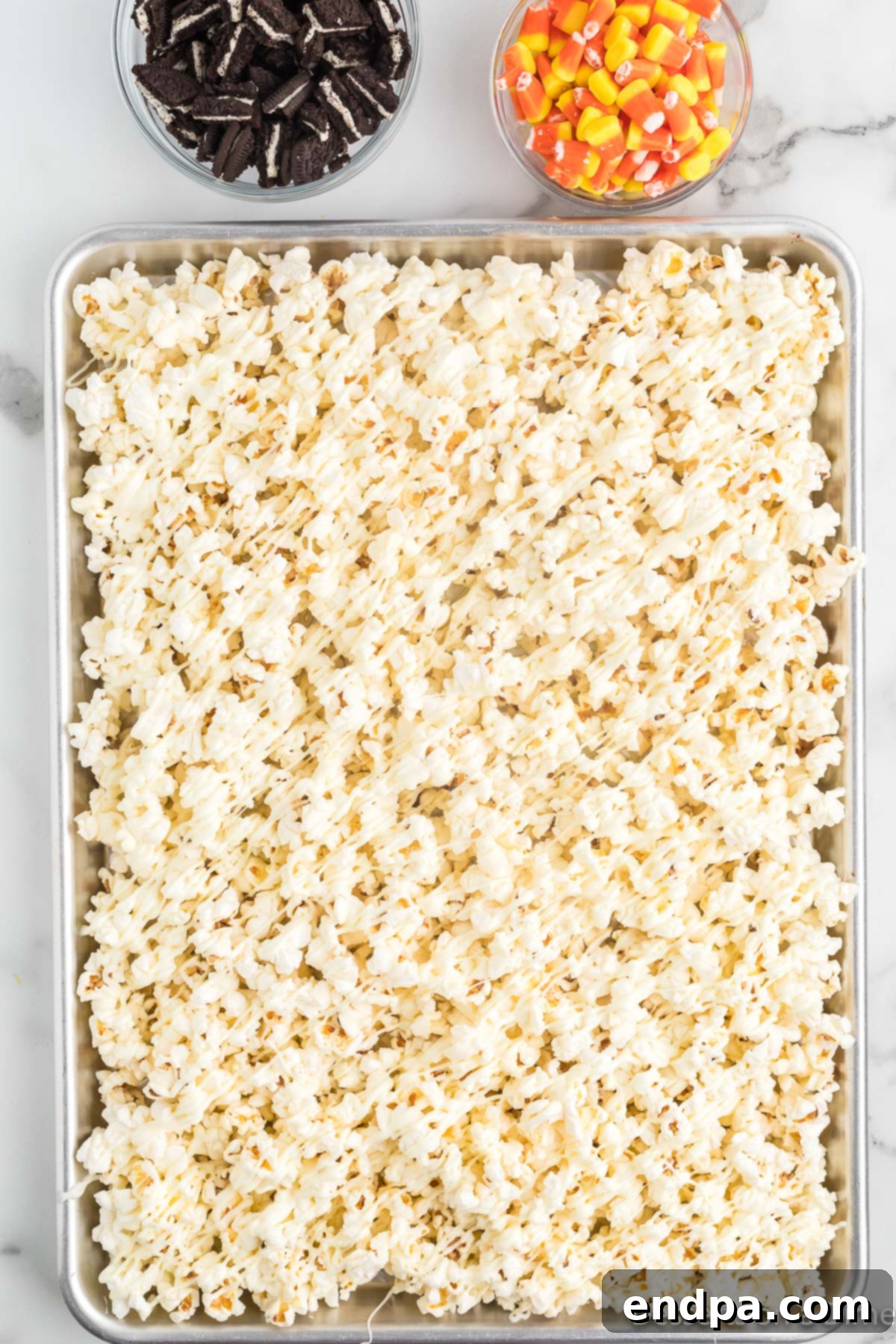 Haunted Kernels 6 Melted white chocolate being drizzled generously over the prepared popcorn on the baking sheet.