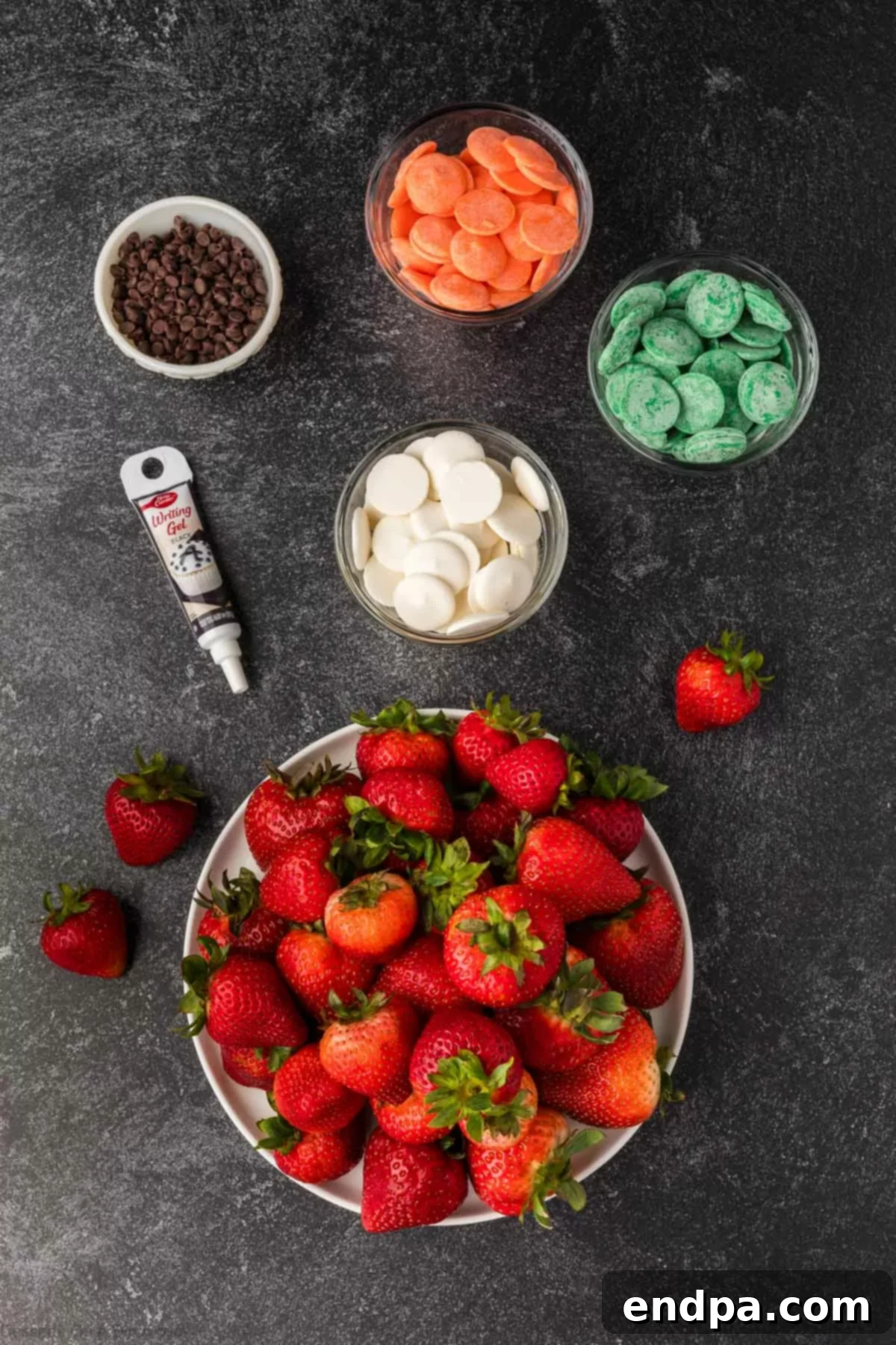A selection of ingredients for Halloween chocolate covered strawberries including fresh strawberries, orange, white, purple, and green candy wafers, black gel icing, and mini milk chocolate chips on a black surface.
