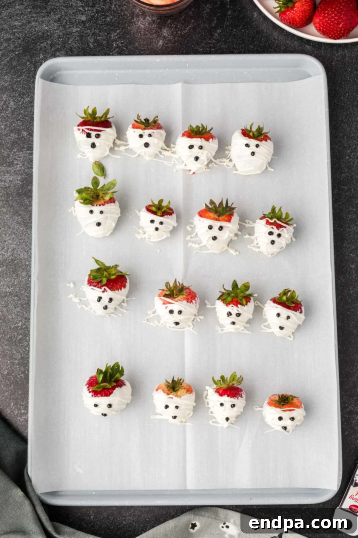 White chocolate covered strawberries on a baking sheet, some decorated with black gel icing faces, resembling ghosts and mummies.