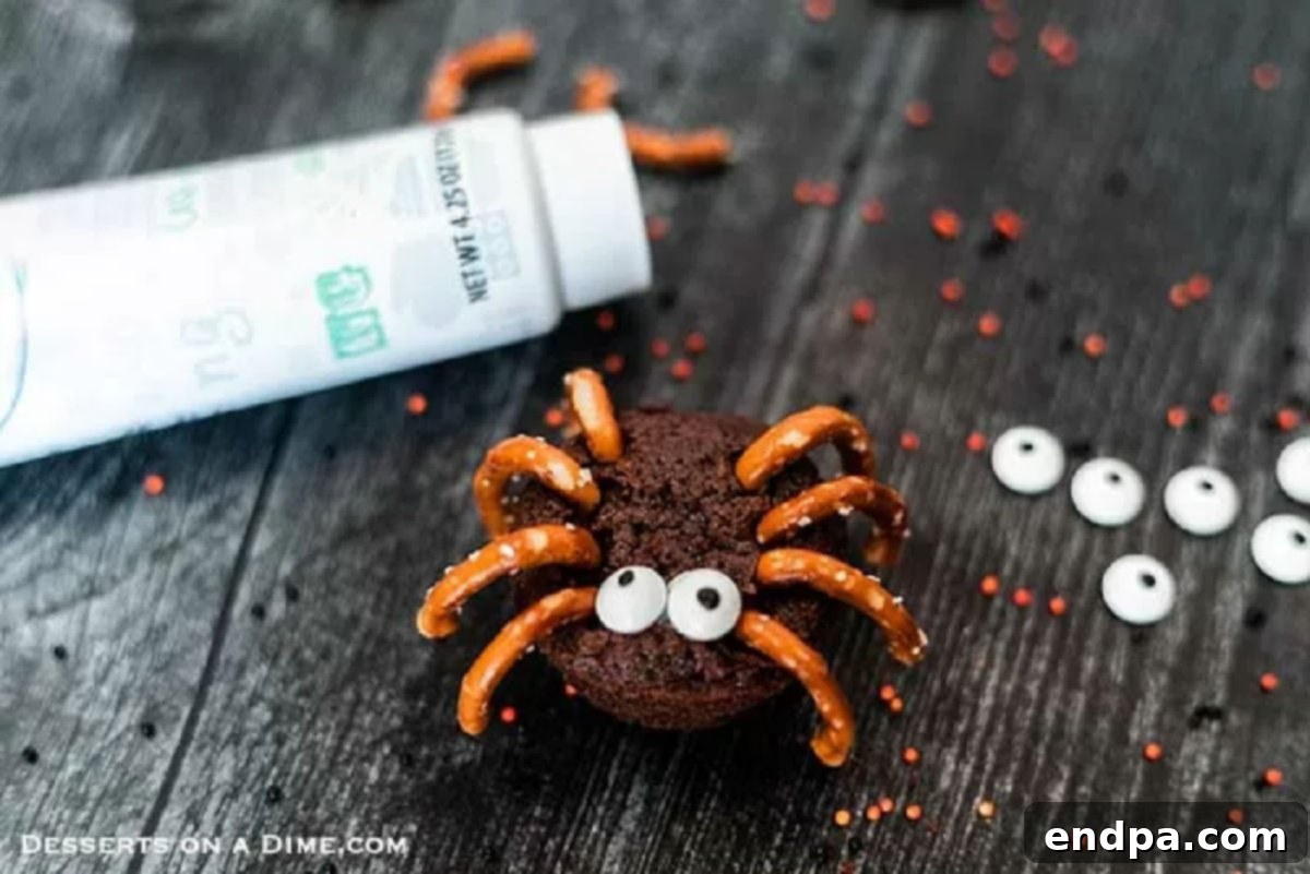 Applying white icing to the front of a mini brownie to attach vibrant candy eyes, completing the adorable spider design.