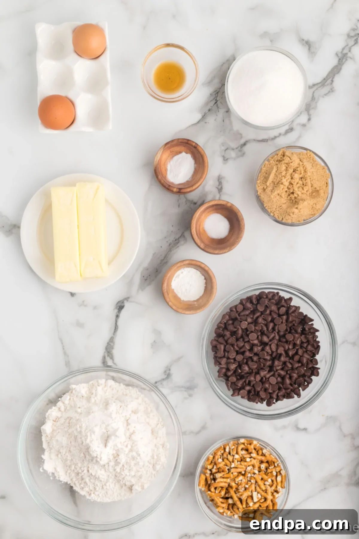 A selection of ingredients for Chocolate Chip Pretzel Cookies, including butter, brown sugar, eggs, vanilla, flour, baking soda, pretzels, and chocolate chips.