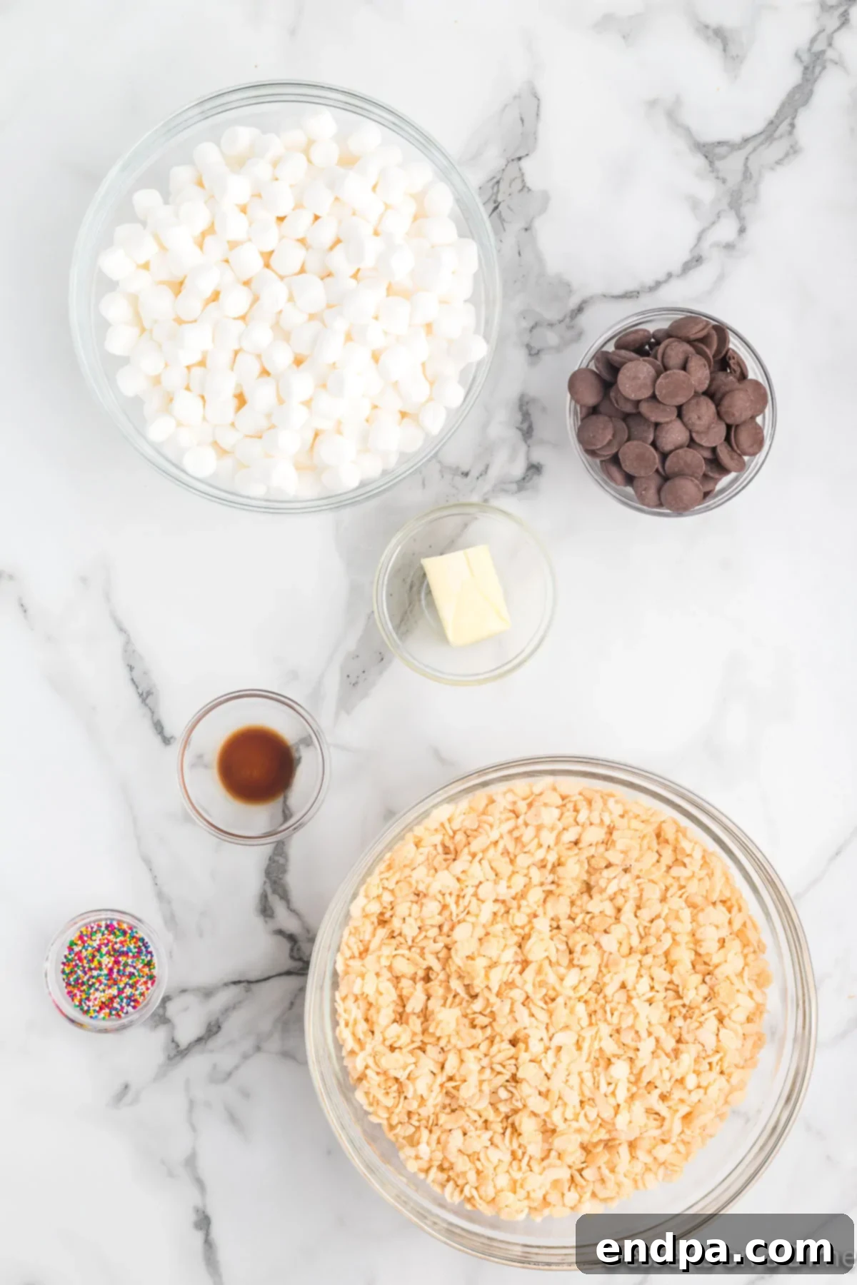 A display of ingredients for Chocolate Covered Rice Krispie Treats: Rice Krispies cereal, butter, mini marshmallows, vanilla extract, chocolate melting chips, and colorful sprinkles.