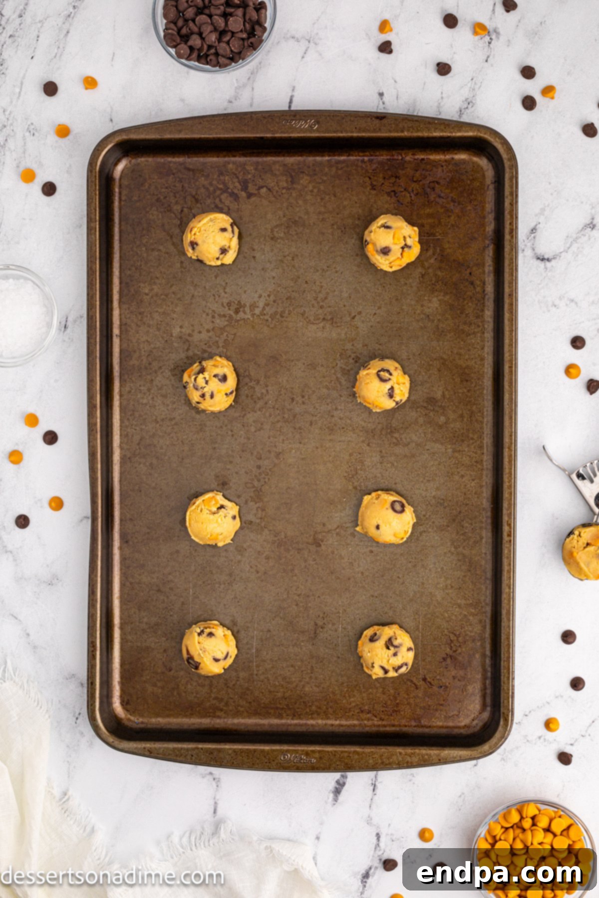 Scoops of cookie dough arranged neatly on a baking sheet lined with parchment paper, topped with extra chips and sea salt.