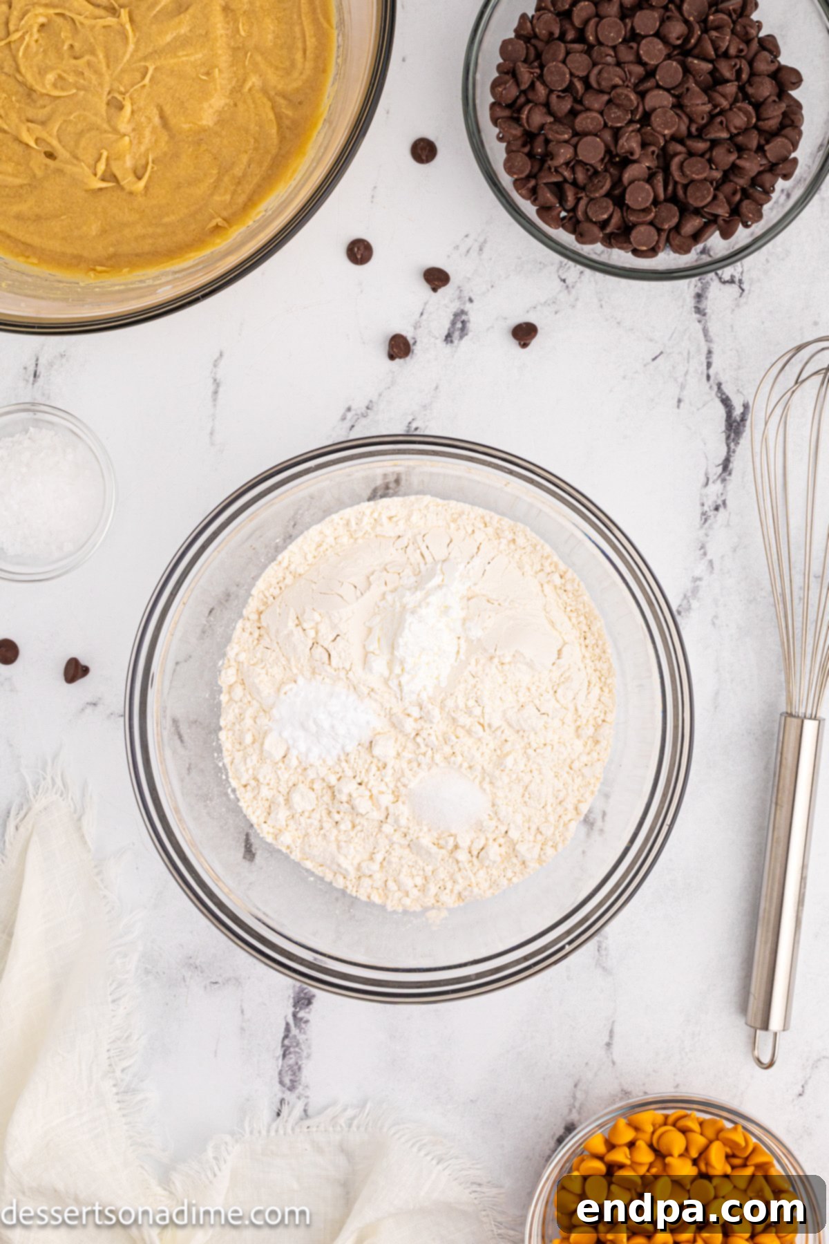 A collection of ingredients for Butterscotch Chocolate Chip Cookies, including butter, light brown sugar, granulated sugar, eggs, vanilla extract, flour, baking soda, cornstarch, salt, chocolate chips, and butterscotch chips.