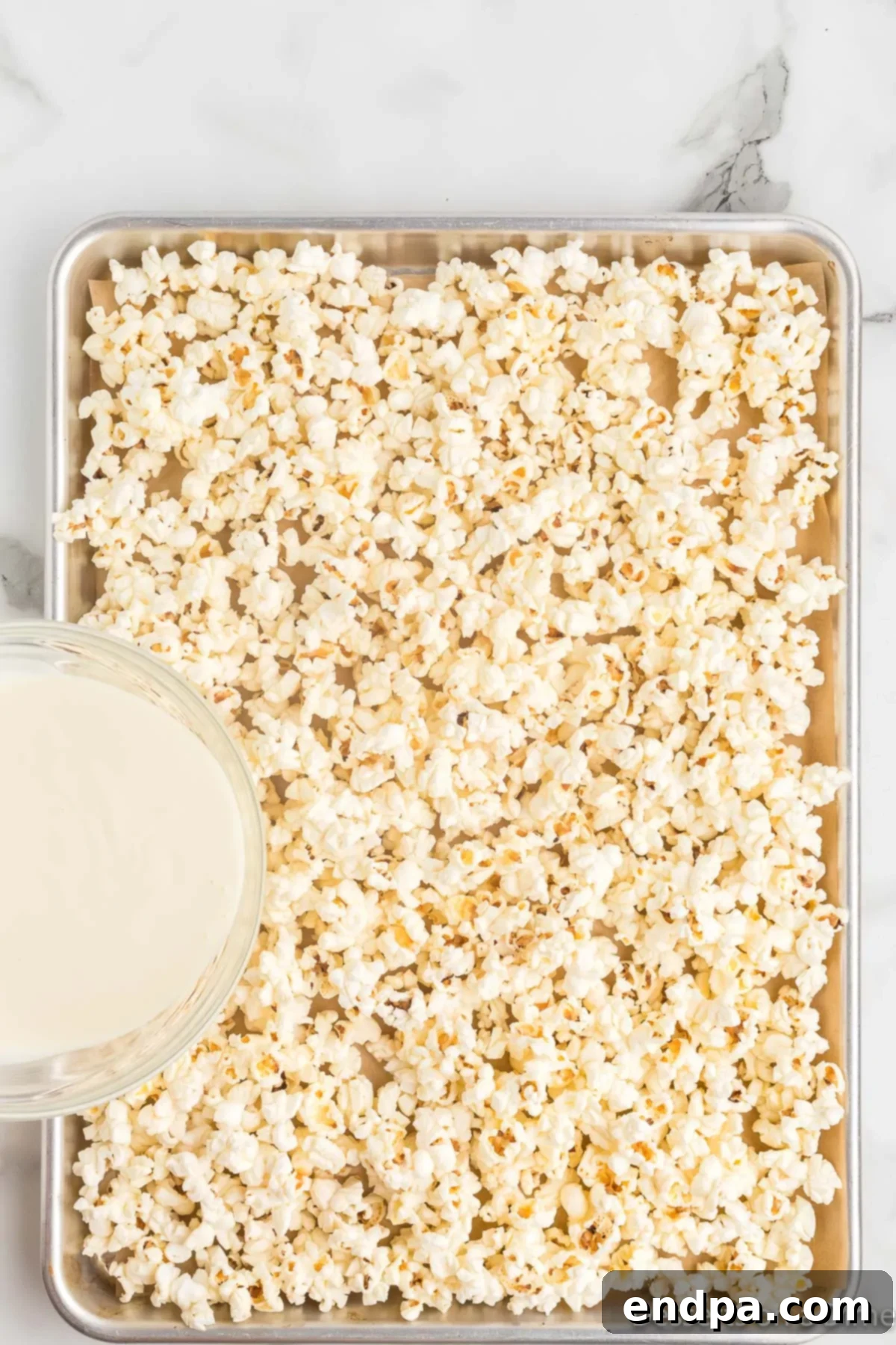 A baking sheet covered with parchment paper and a generous layer of freshly popped popcorn, ready for the white chocolate drizzle.