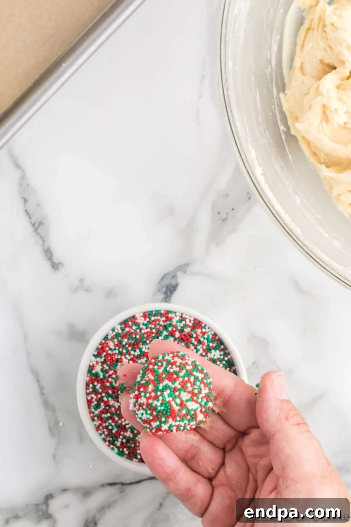 A dough ball being thoroughly rolled in a shallow bowl of festive red, green, and white sprinkles until completely covered.
