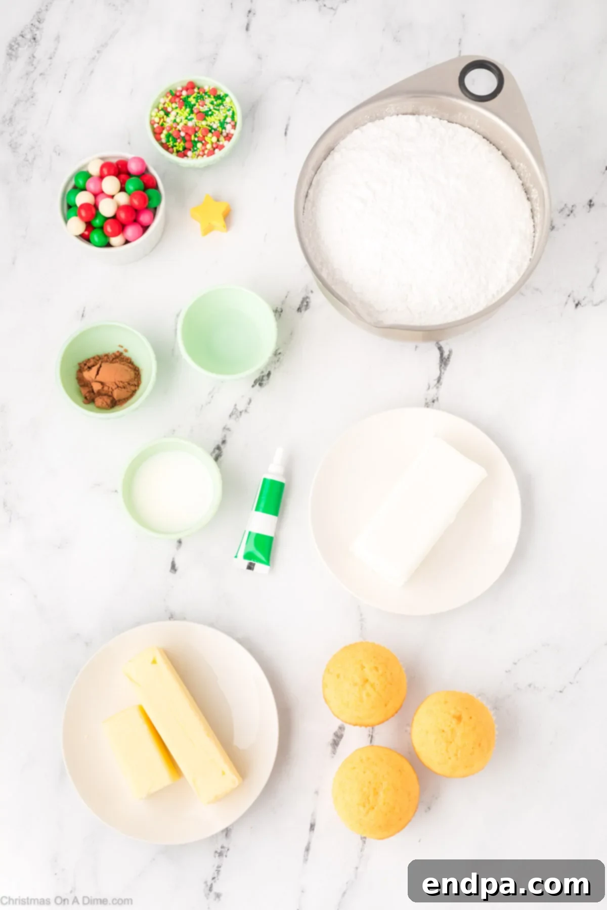 A flat lay image displaying all the necessary ingredients for Christmas Tree Cupcakes: vanilla cupcakes, butter, clear vanilla extract, powdered sugar, milk, green gel food coloring, cocoa powder, and festive sprinkles.