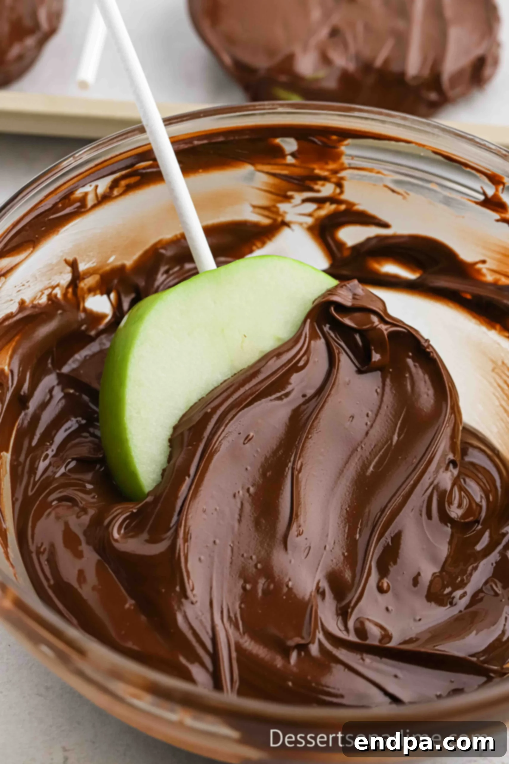 Melted chocolate in a bowl with an apple slice being coated.