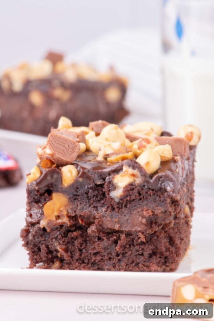 Snickers Overload Cake 2 A slice of Snickers cake on a white plate, showcasing layers of chocolate cake, caramel, peanuts, and chocolate frosting.