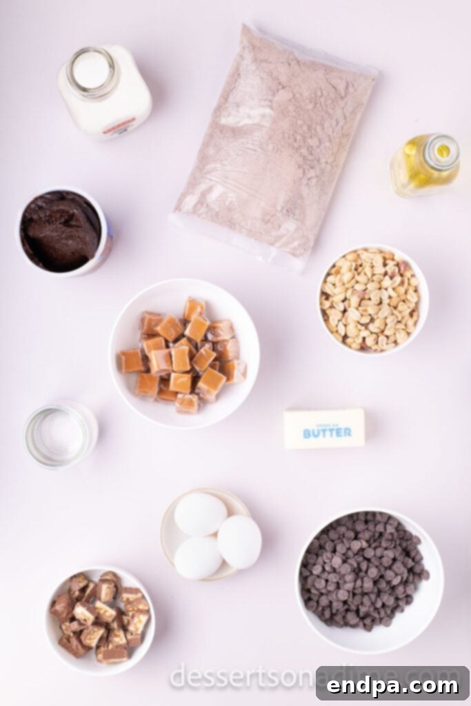 Snickers Overload Cake 3 A flat lay of ingredients for Snickers Cake including a box of cake mix, eggs, butter, oil, caramel candies, chocolate chips, and whole Snickers bars.