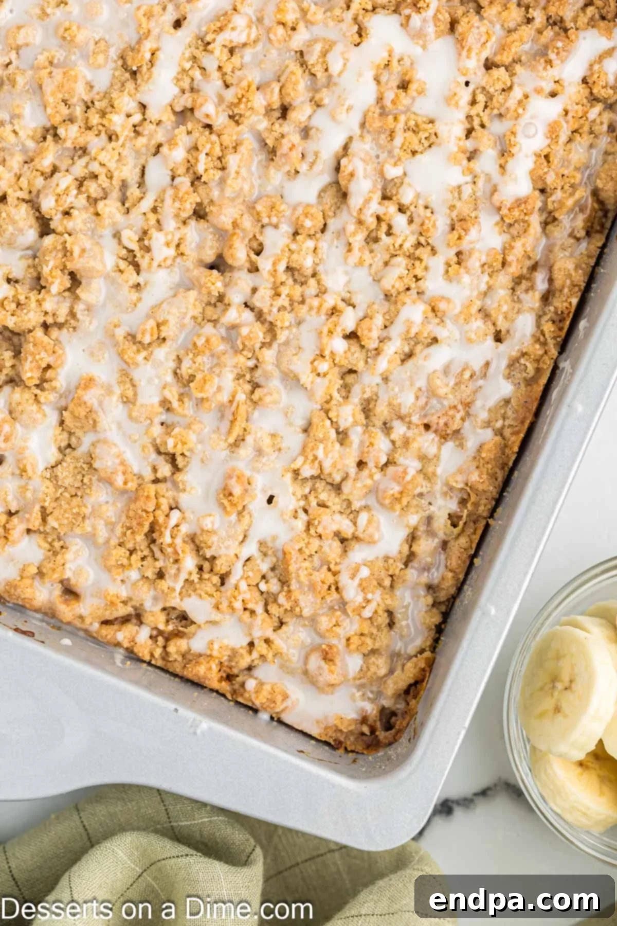 A freshly baked banana coffee cake in its rectangular pan, complete with a golden streusel topping and delicate vanilla glaze, ready for slicing and serving.