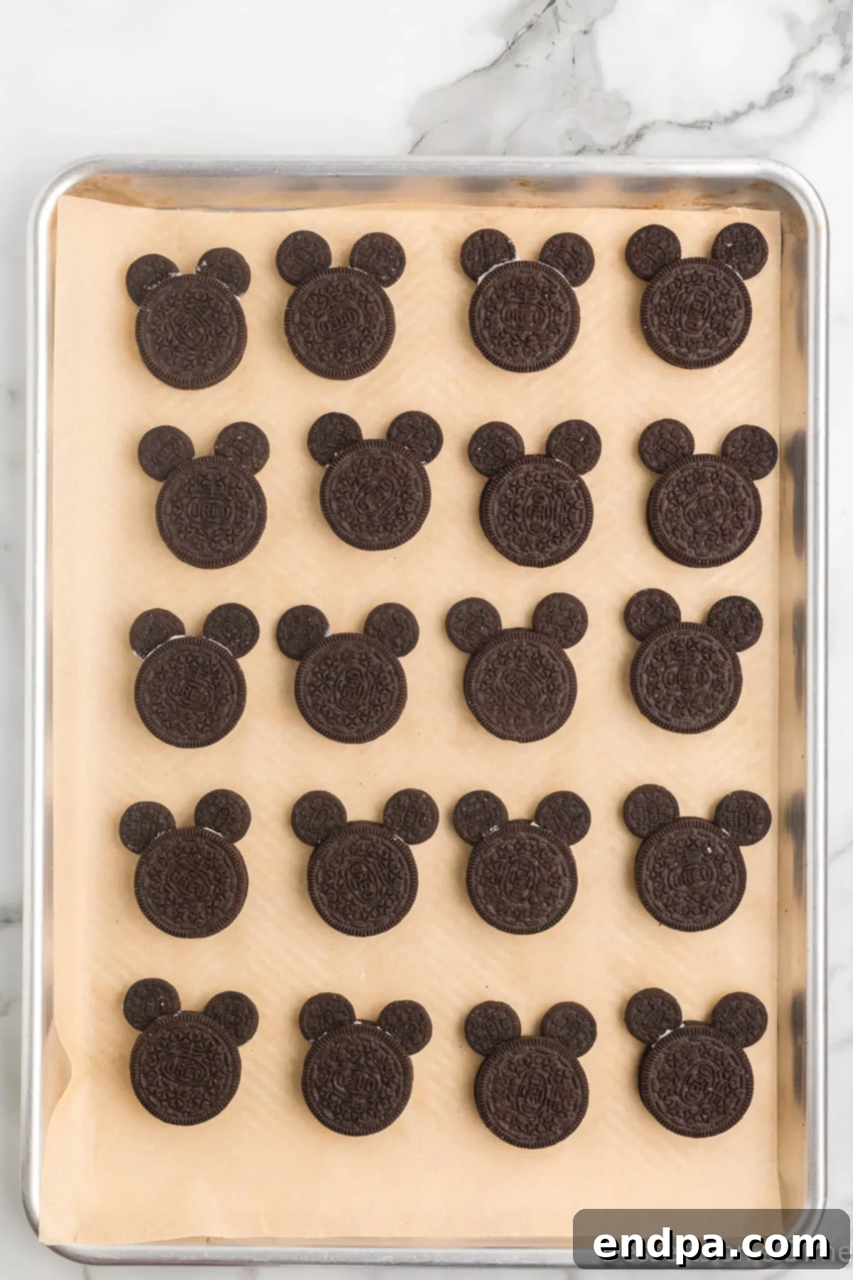 Mickey's Magical Oreo Treats 6 A double-stuffed Oreo cookie on a baking sheet, with two mini Oreo halves carefully inserted into the cream filling to form Mickey Mouse ears, prior to dipping.