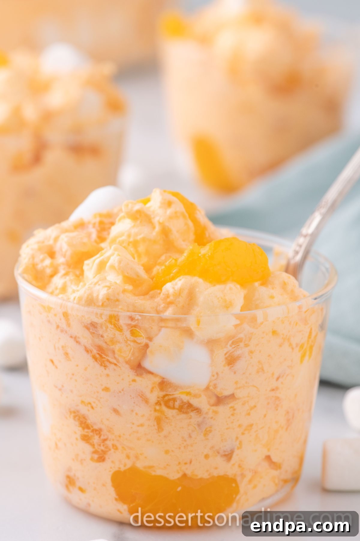A serving of creamy Orange Fluff in a clear glass cup, garnished with a spoon, highlighting its vibrant color and fluffy texture.