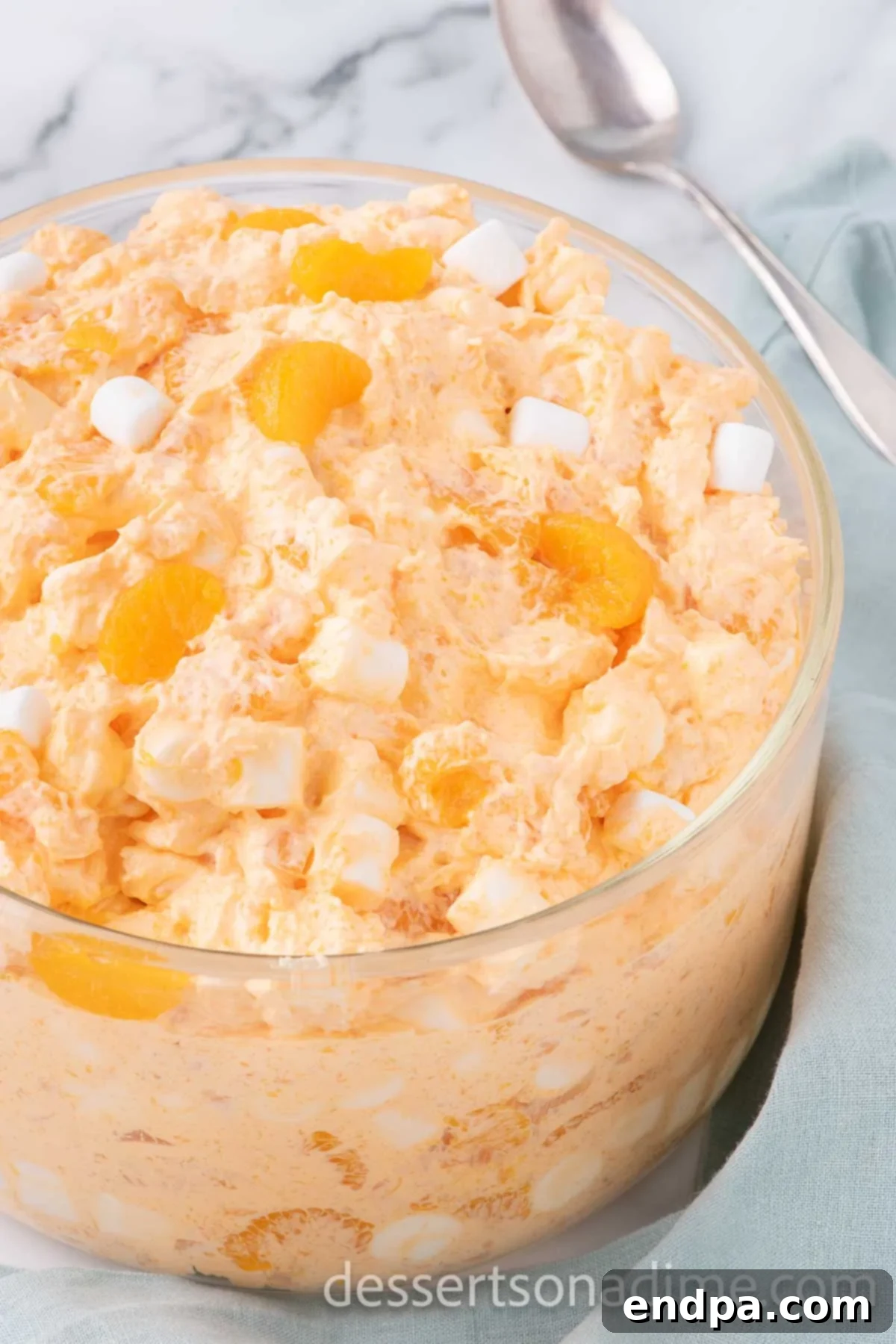 A large bowl of vibrant Orange Fluff covered tightly with plastic wrap, ready to be placed in the refrigerator for its final chilling phase.