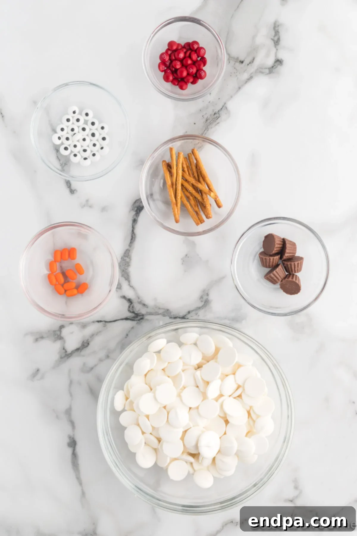 Frosty Meltdown Bark 3 Ingredients for Melted Snowman Bark: candy eyes, pretzel sticks, orange Tic Tacs, mini Reese's cups, and white candy melts.