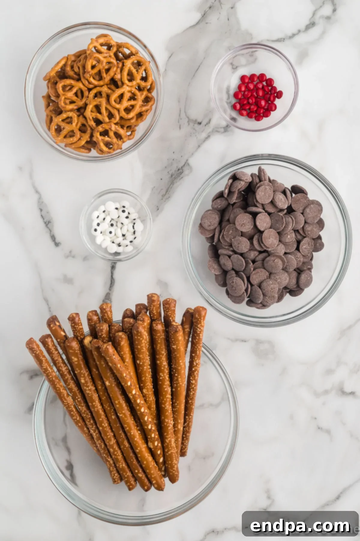 Ingredients for Reindeer Pretzel Rods including large pretzel rods, candy melting chocolate, candy eyes, mini red M&Ms, and pretzel twists.