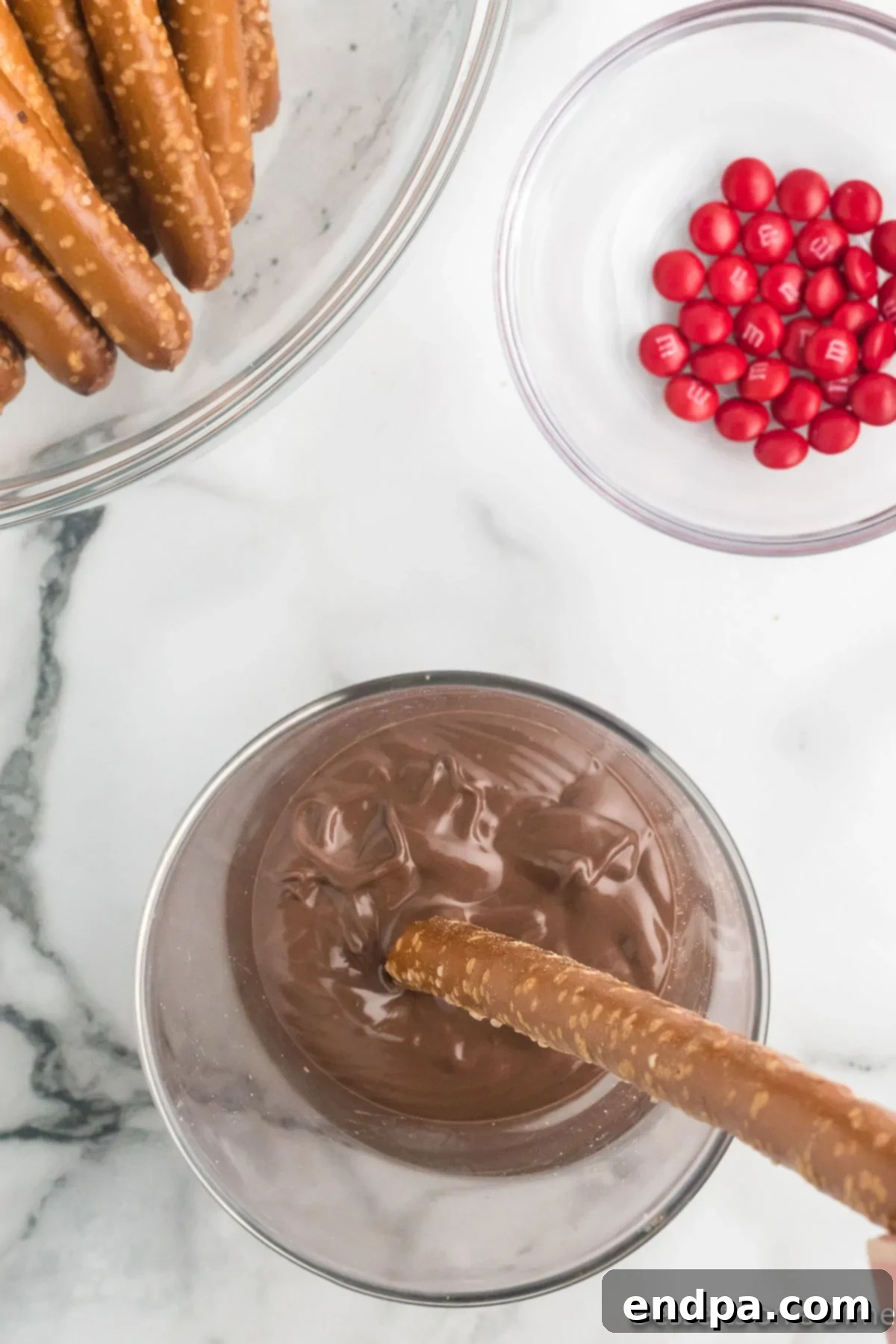 A large pretzel rod being carefully dipped into a bowl of melted chocolate, covering approximately half of its length.