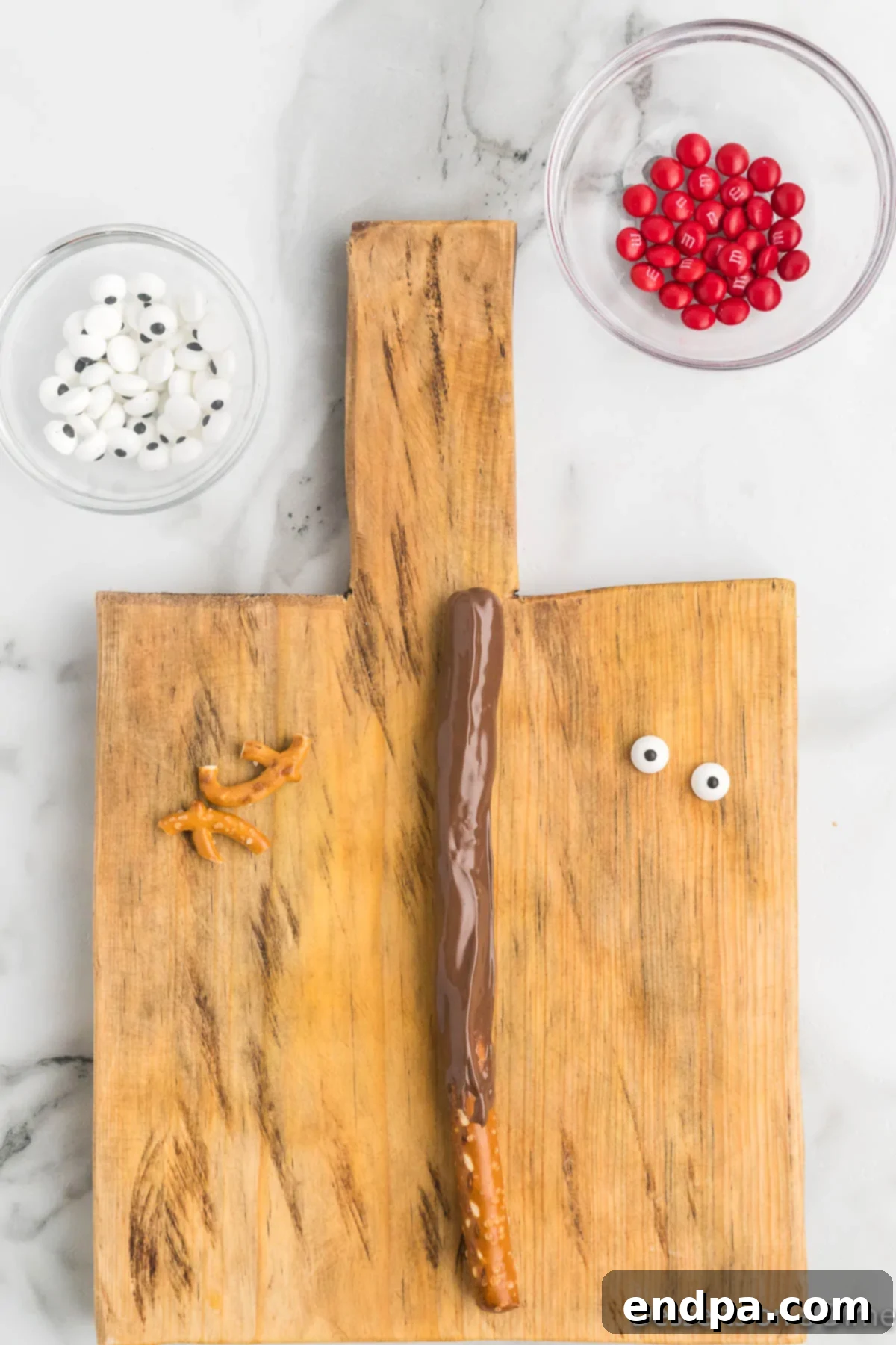 Various candy components for reindeer faces – candy eyes, red M&Ms, and pretzel antler pieces – arranged on a surface.