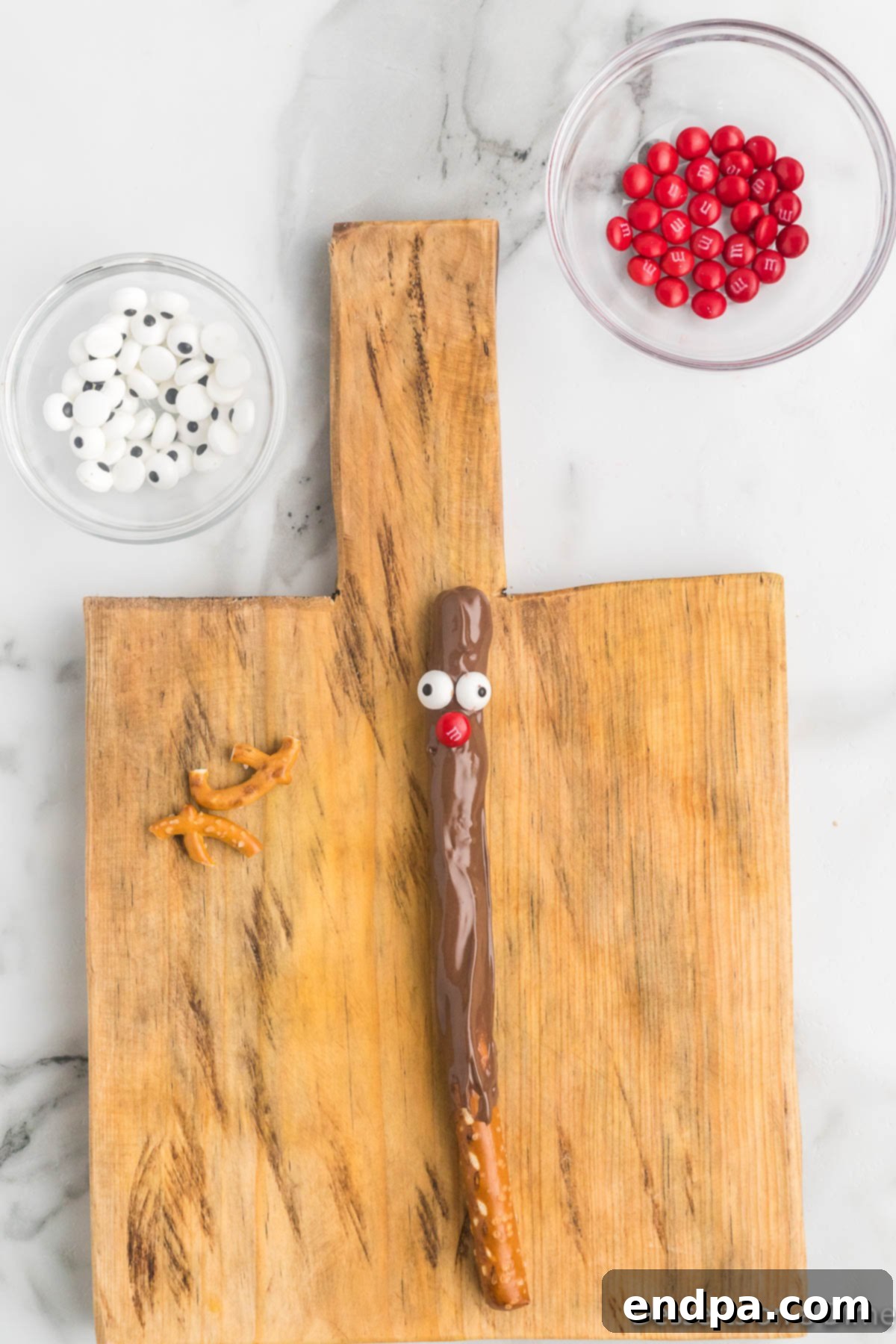 Two candy eyes and a red M&M being placed onto the wet chocolate of a dipped pretzel rod to form a reindeer face.