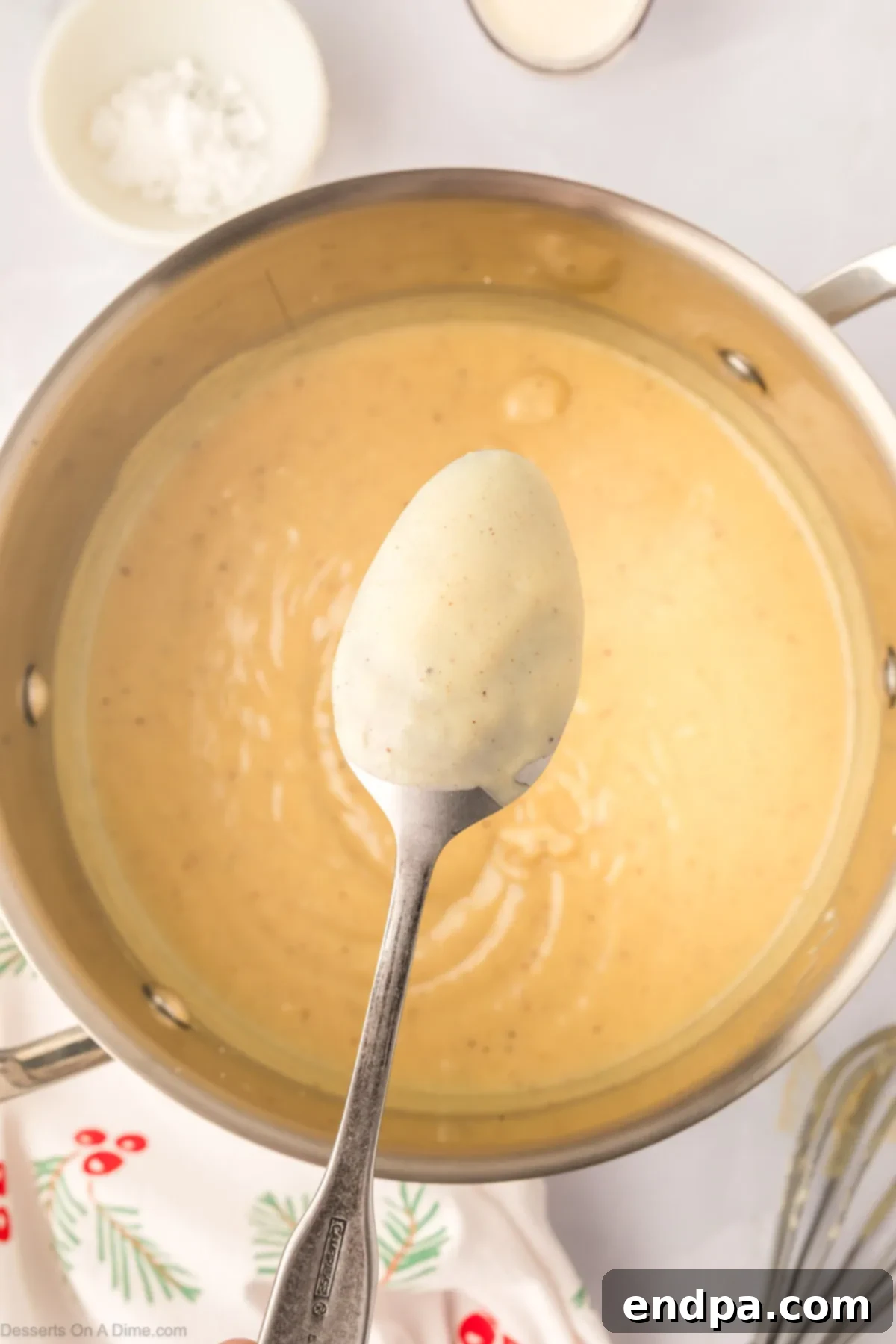 A whisk lifted from the thickening eggnog pudding on the stovetop, demonstrating the perfect consistency where the mixture coats the back of a spoon.